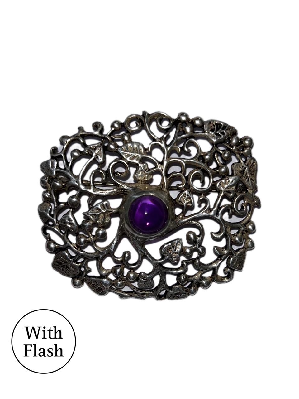 This beautiful brooch is floral-themed. The silver-toned brooch is a rectangle made of intertwined leaves and branches. A purple round cabochon is placed at the centre, which reflects the light beautifully and brings a nice touch of colour. On the