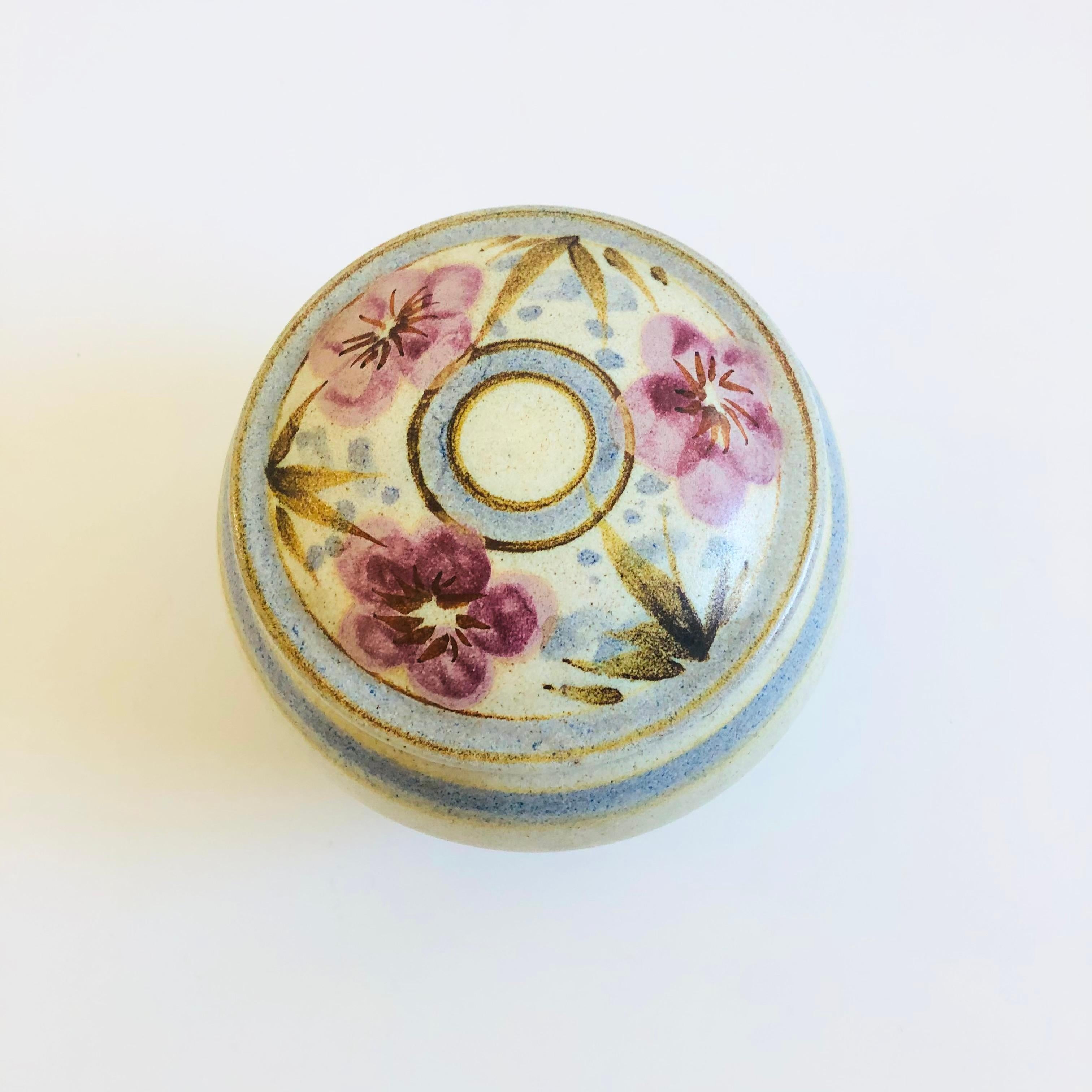 A vintage handmade studio pottery lidded container. Nice round shape with a speckled gray base glaze. Bands of blue wrap around the sides with a lovely hand painted floral design to the lid. Signed by the artist, Leila, on the base.

