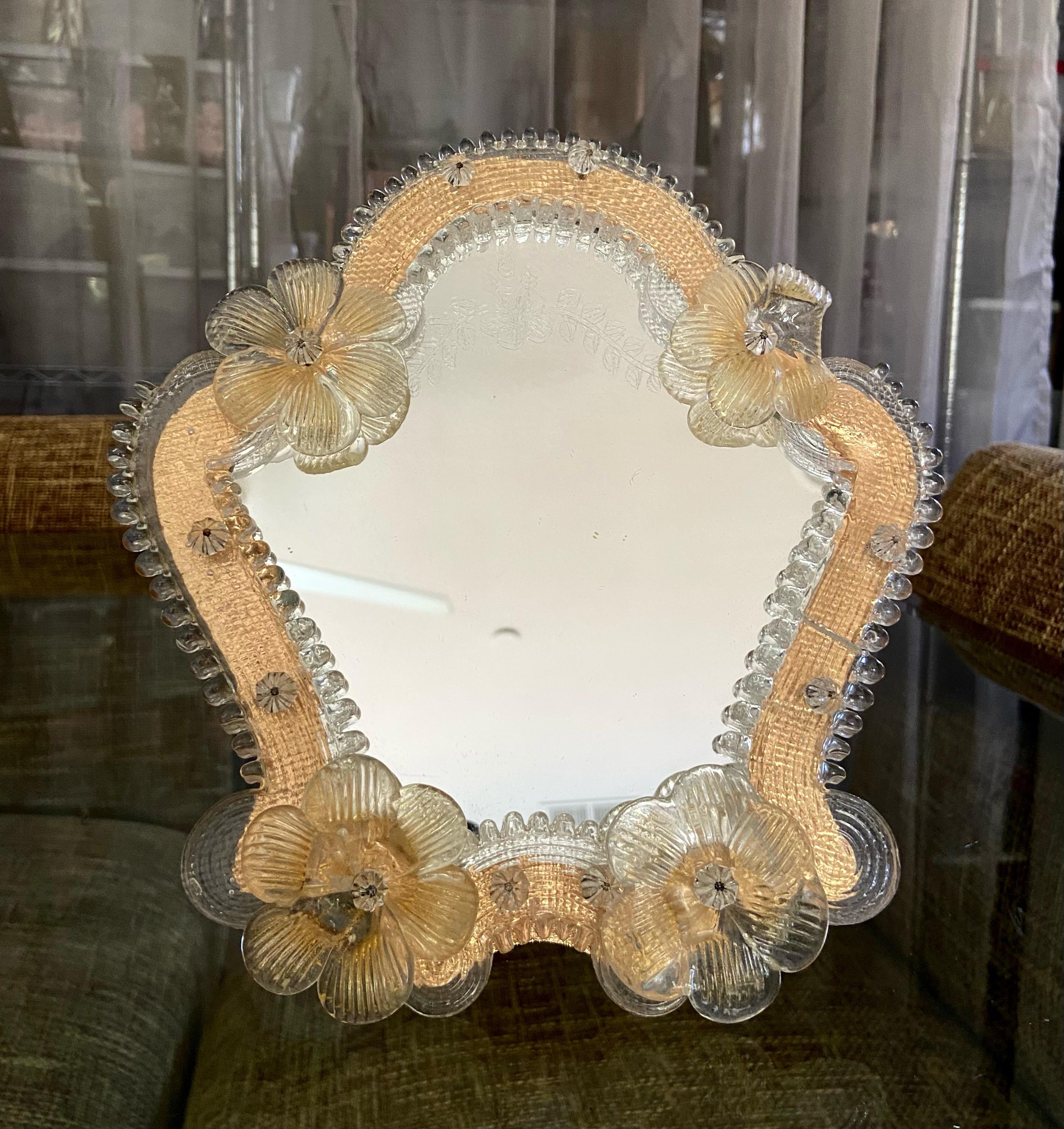 Kleinerer Muranospiegel mit klaren und goldenen Blumen und hellen bernsteinfarbenen Glasstücken, die den Spiegel umgeben. Das Spiegelglas hat eine dekorative Ätzung. Der Ständer und die Unterlage sind aus Holz. 
  