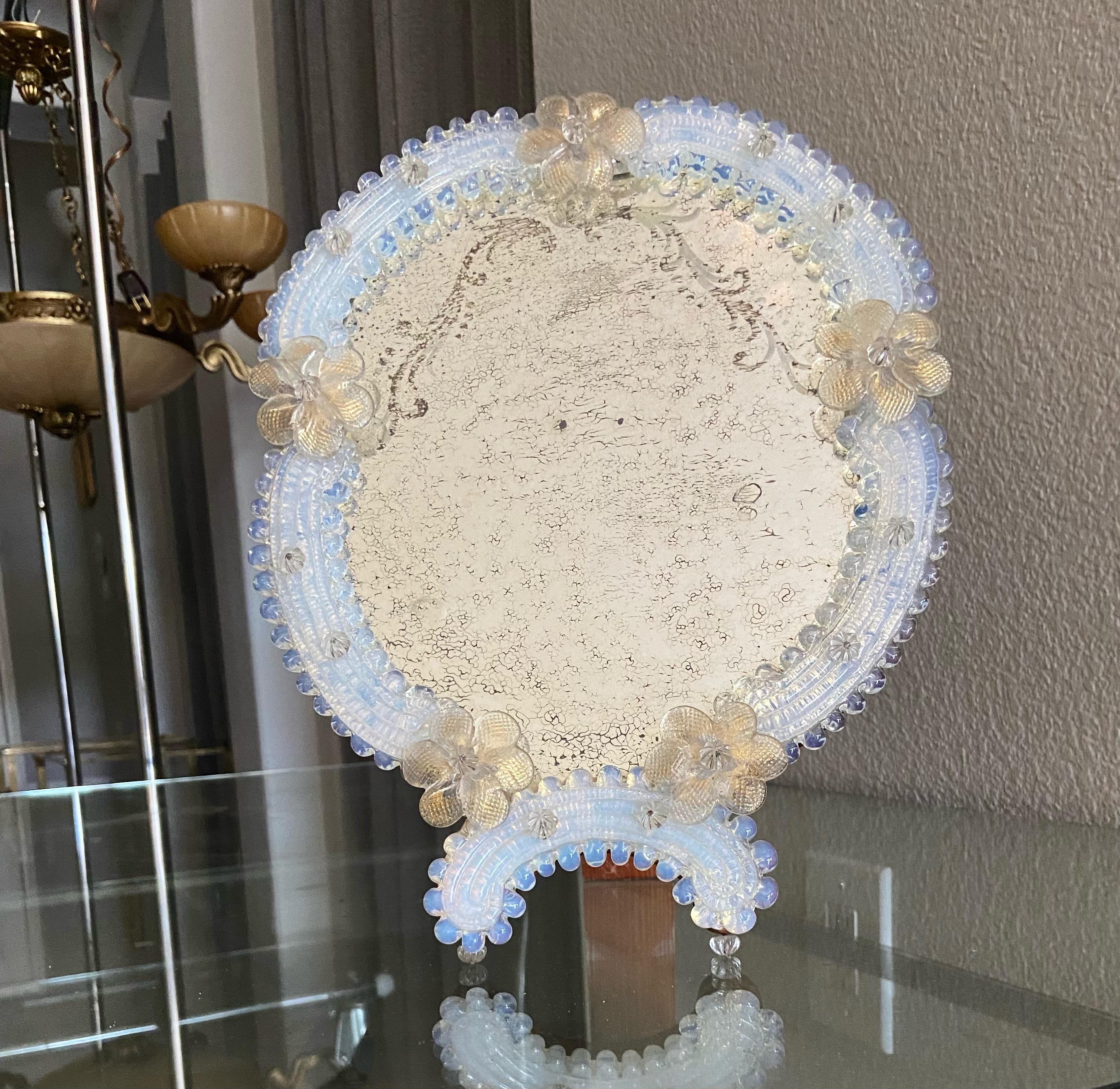 Miroir de table de Murano avec des fleurs transparentes et dorées, et des pièces de verre de couleur opalescente entourant le miroir. Le verre du miroir présente une gravure décorative et une finition antiquaire. Le pied et le support sont en bois.