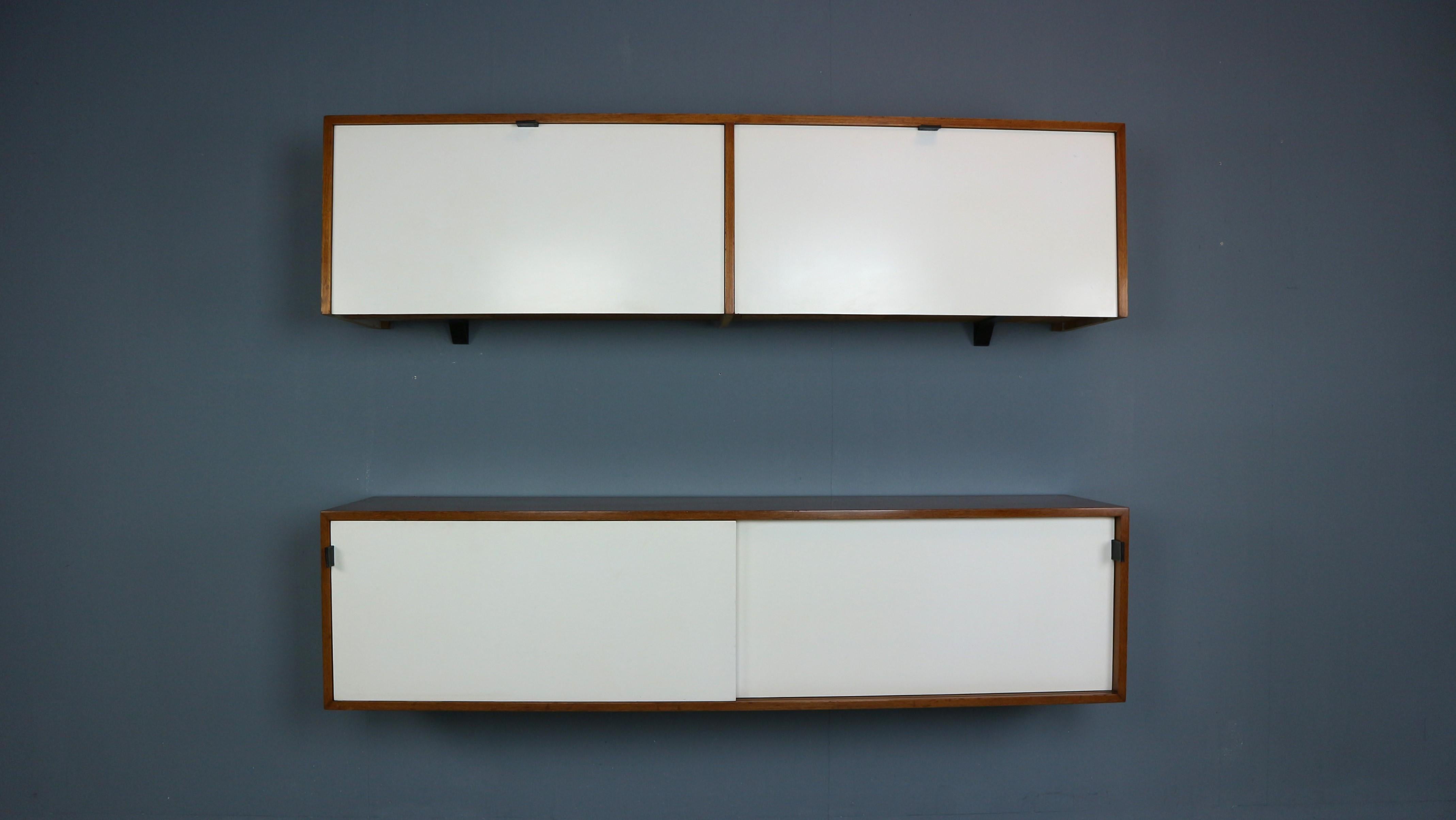 Two walnut and formica wall mount storage cabinets or credenzas designed by Florence Knoll for Knoll Associates. Early production in beautiful walnut wood with white doors, leather/brass handles, interiors and shelves. One cabinet with drop down