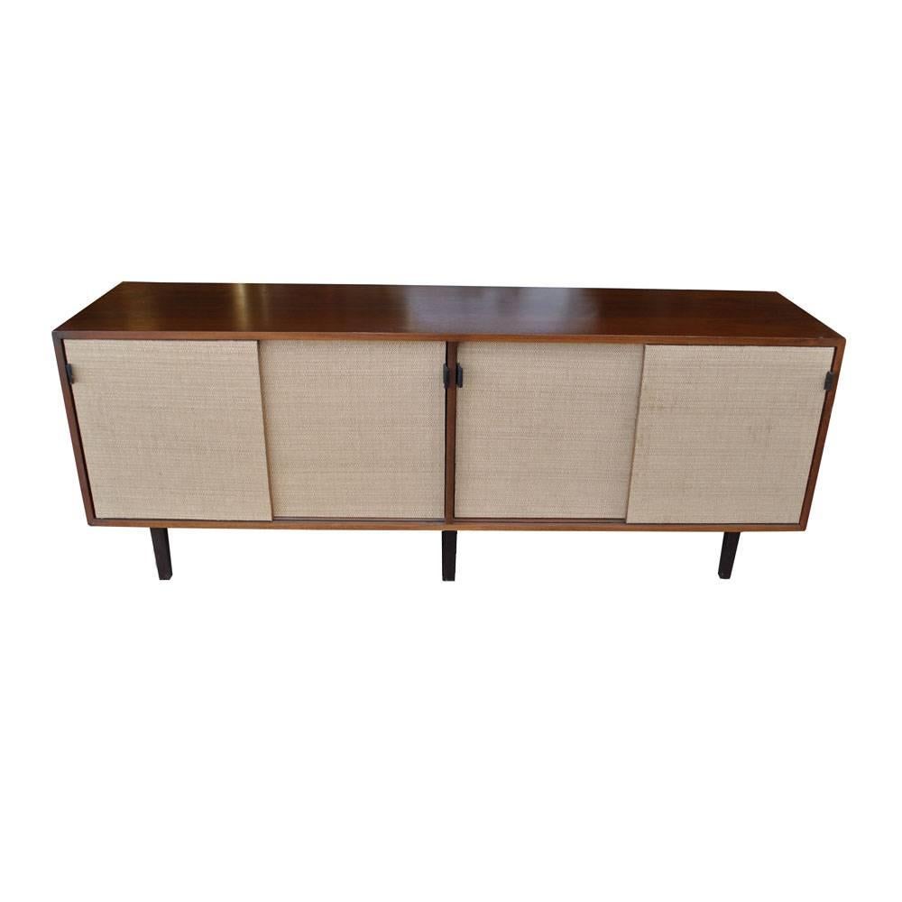 A mid century modern buffet or credenza designed by Florence Knoll and made by Knoll. A walnut case with sliding grasscloth covered doors, leather pulls, and wooden legs. The interior has shelved storage.