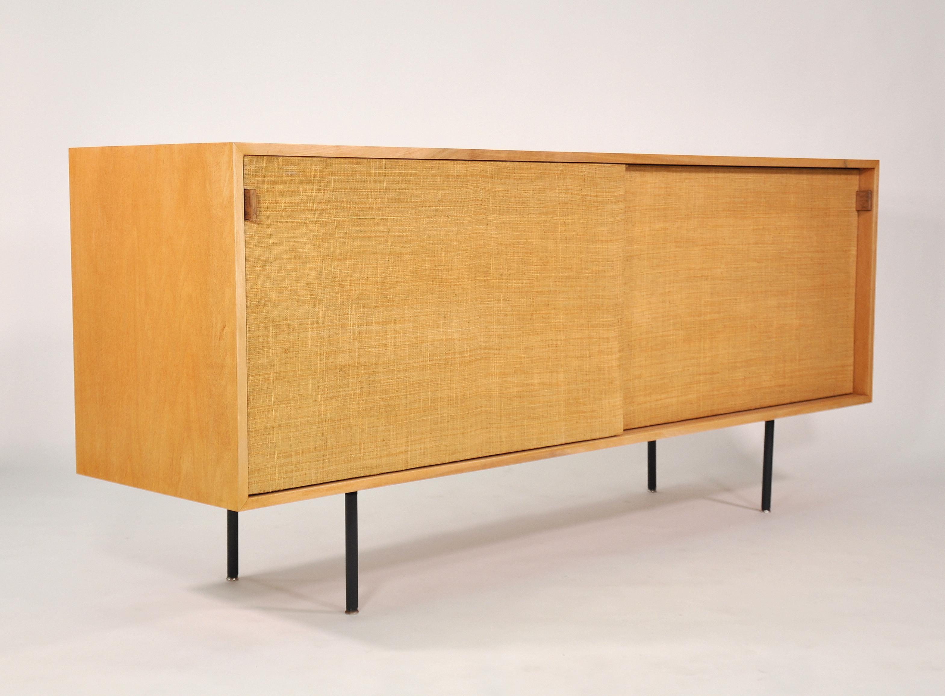 A maple, grasscloth and leather cabinet in the style of the 116 model designed by Florence Knoll in 1948 for Knoll Associates. The sideboard or bar features a pair of sliding doors with seagrass fronts and saddle leather handles, opening to a white