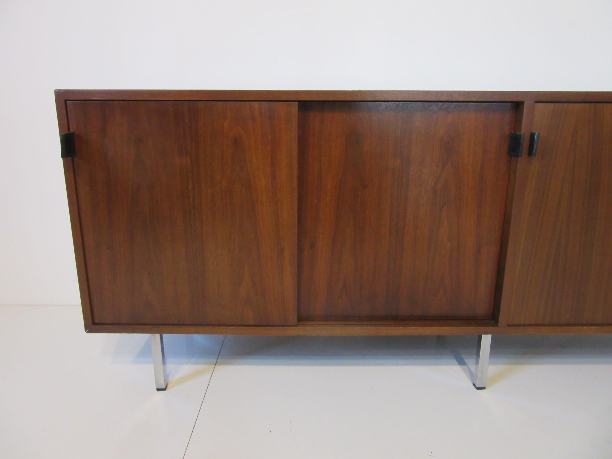 A well-constructed dark walnut credenza with four sliding doors, black leather pulls, two storage areas and two areas with adjustable shelves sitting on square chromed legs. The inside has a natural oak contrasting finish with painted satin black