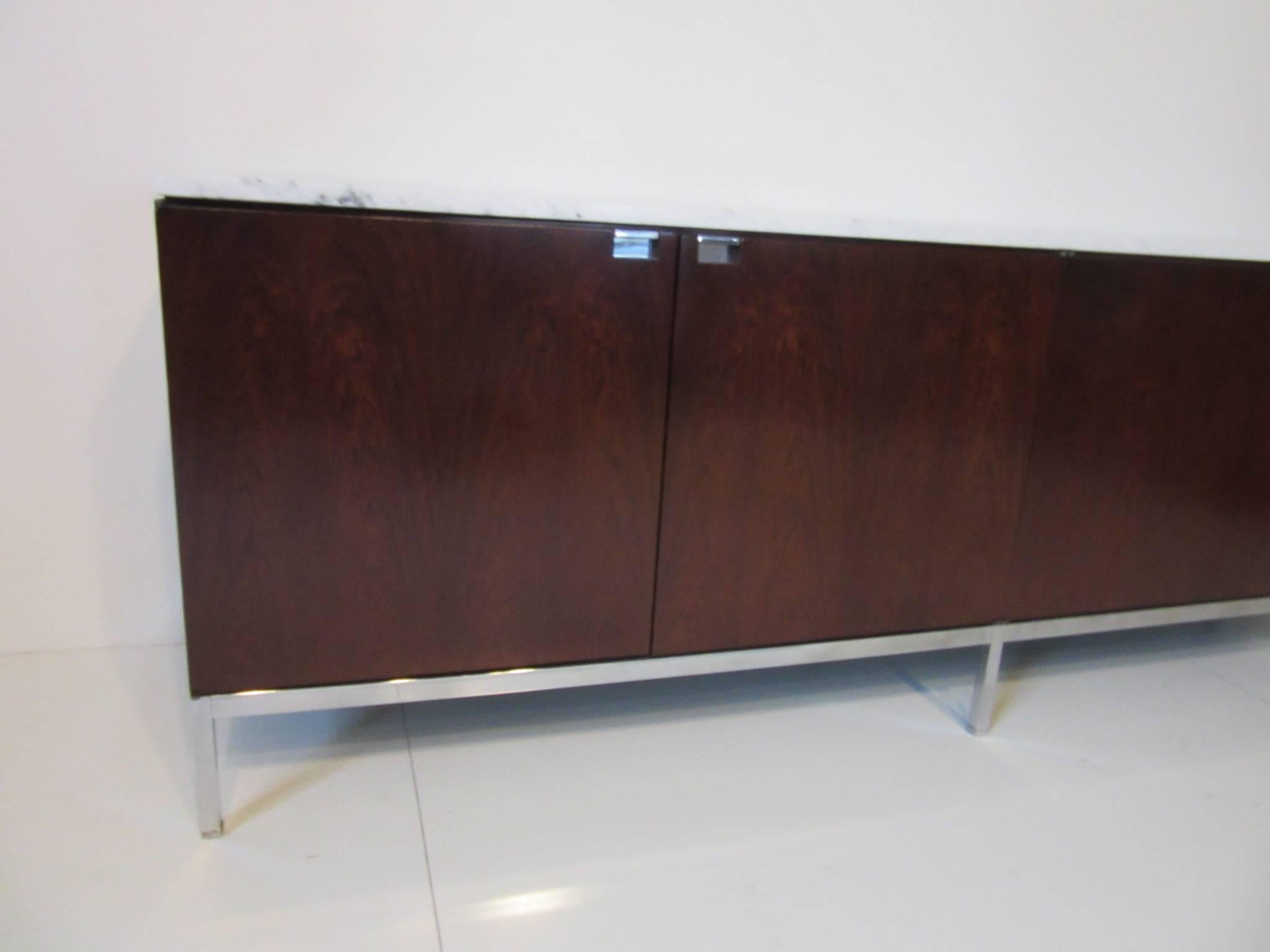 A dark Brazilian rosewood credenza with white Carrara marble top, four doors, adjustable shelves, chrome pulls, bottom frame and legs. Manufactured by Knoll International.