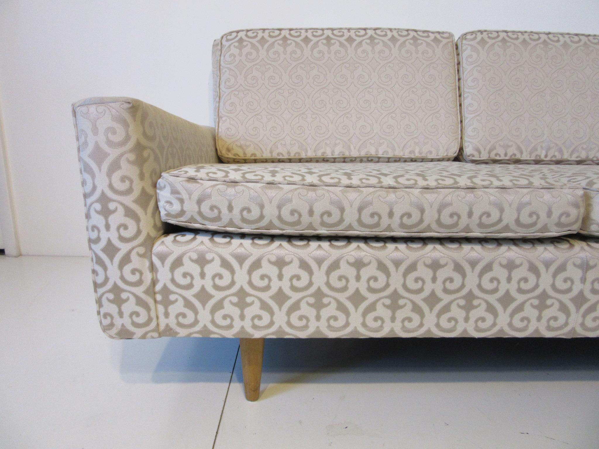 A two cushion settee / loveseat with inset clear birch conical legs , angled arms and side profile upholstered in a Damask fabric with slight shimmering back round . A early production sofa , model # 27 by Knoll .