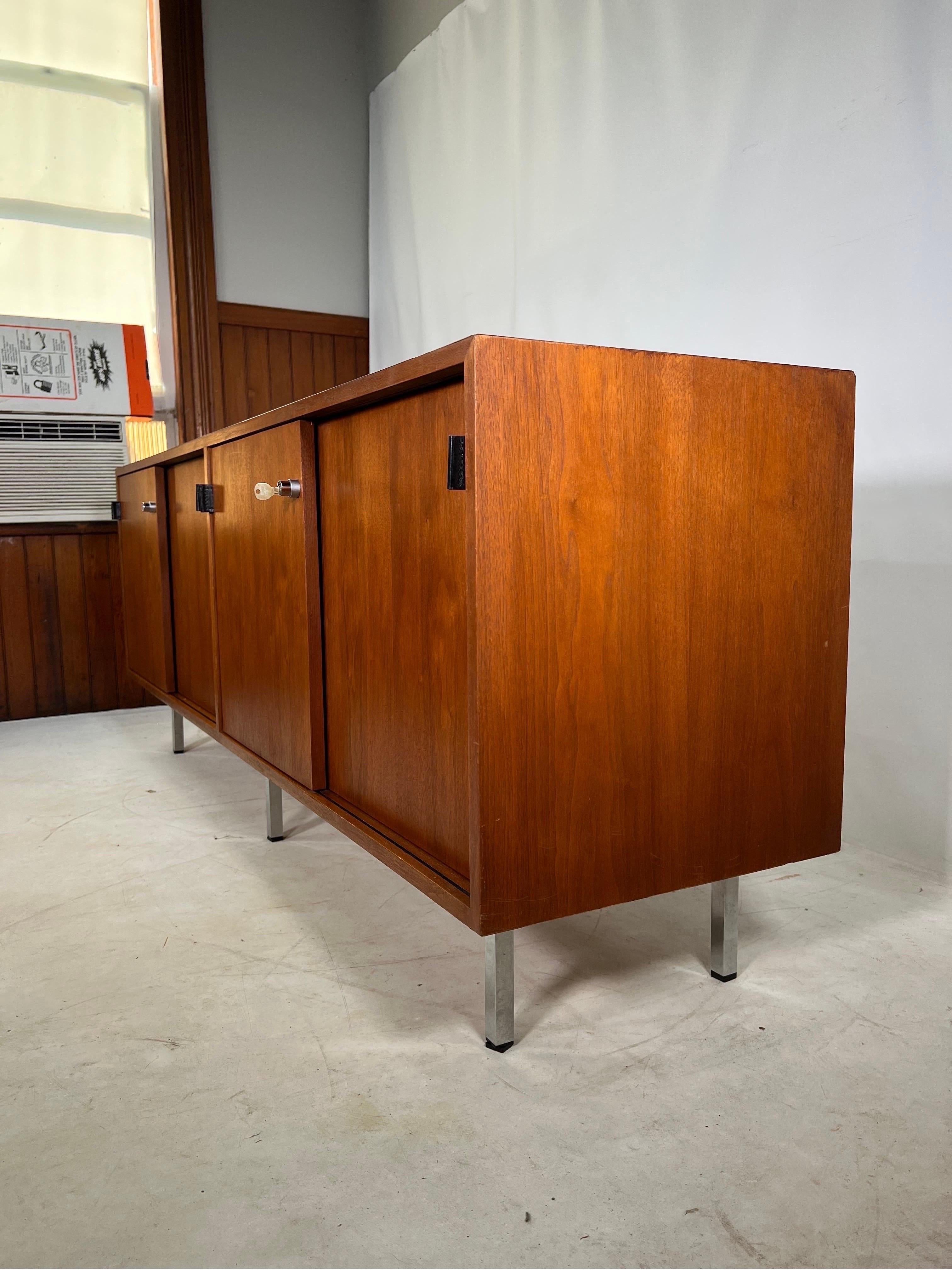 The credenza is constructed of walnut wood front sliding doors, walnut sides, leather strap handles, chrome legs and a laminate top. The credenza still has its original knoll tag and key.
