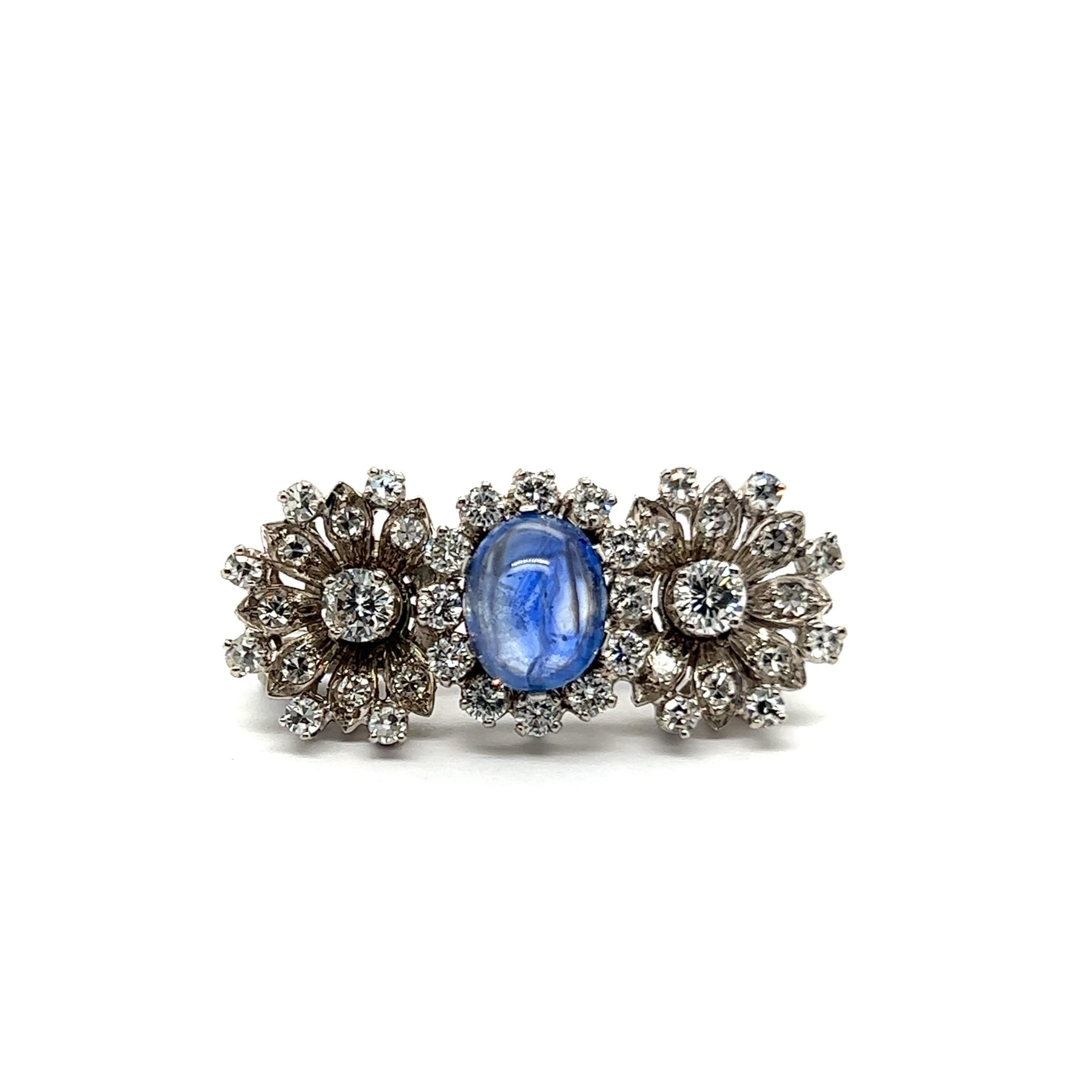 Presenting a delightful flower brooch adorned with blue sapphire and diamonds in 18 Karat white gold. 

At its centers is a 2.80-carat cabochon sapphire encircled by a halo of diamonds. Two accompanying blossoms feature two brilliants in G-H color