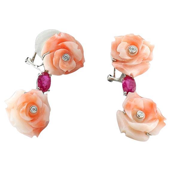 A pair of enchating earrings featuring intricately carved roses from red coral as the central motif. These flowers are complemented by rubies and diamonds, creating an elegant and vibrant design. The coral roses are connected by pink rubies totaling