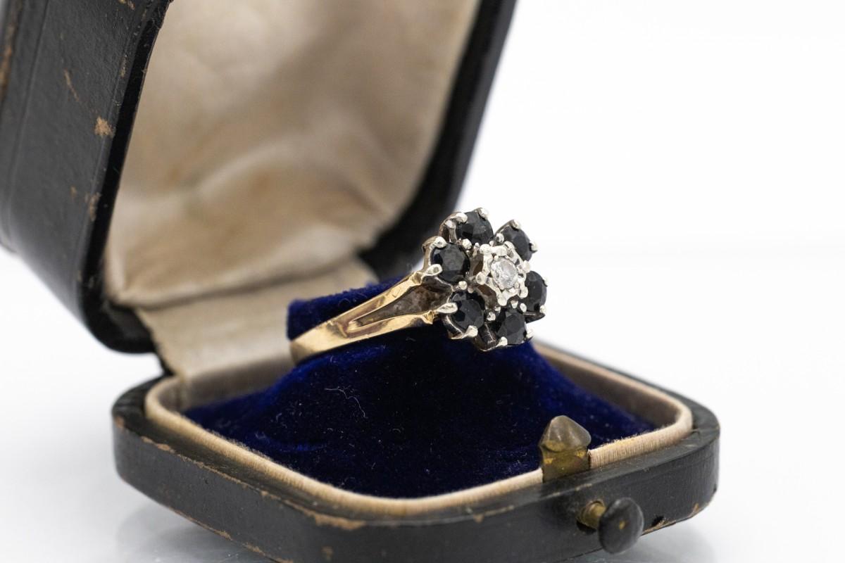 Un affascinante anello d'oro con zaffiri blu scuro e un diamante centrale. Realizzato in oro giallo e bianco a 9 carati. È decorato con 6 zaffiri naturali di colore blu scuro e un peso totale di circa 1,20ct. Il diamante centrale ha un peso di circa