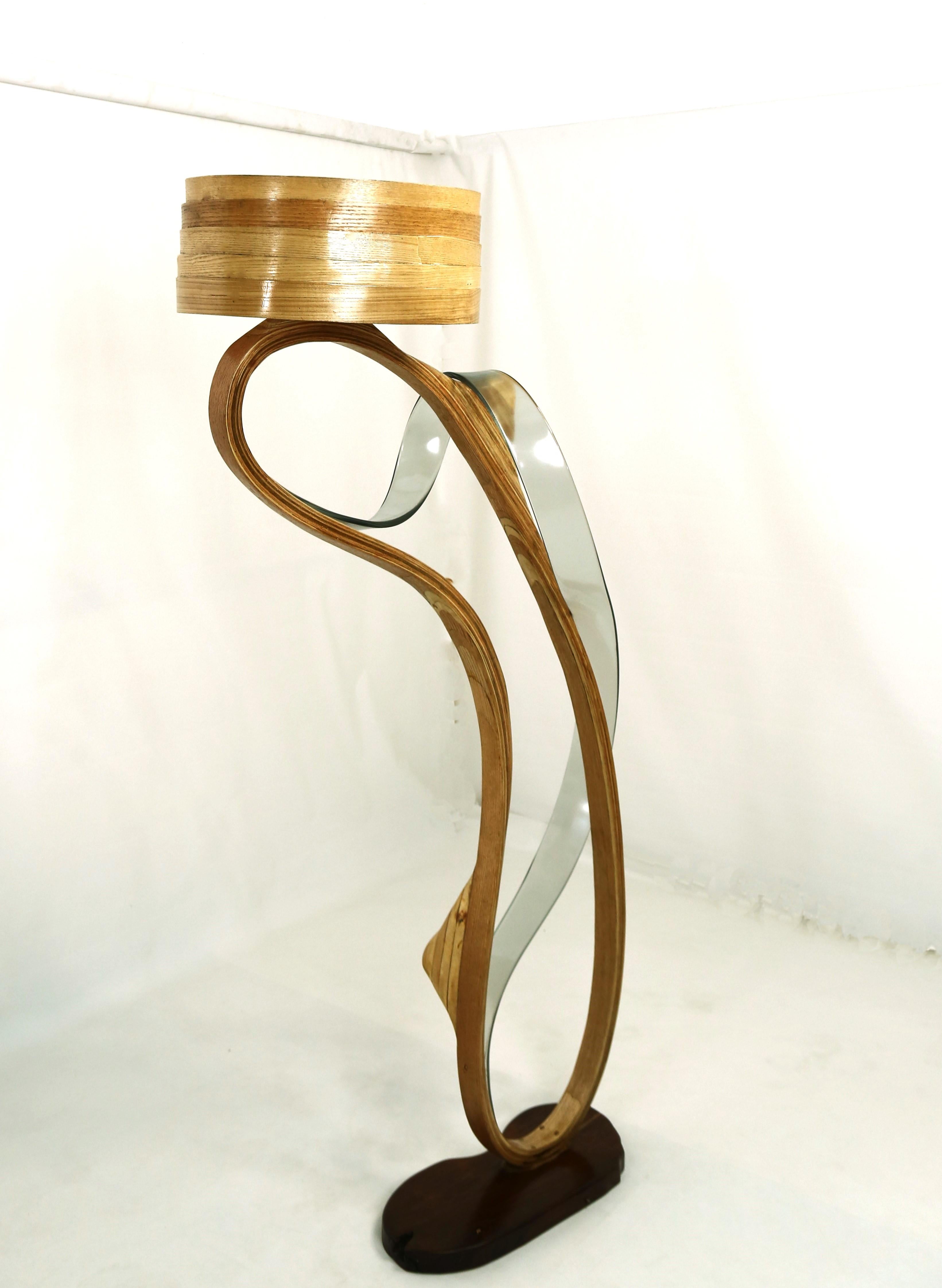 La lampe Fluctus a été fabriquée en bois de frêne avec différentes techniques d'empilage et un design minimal. Une bande transparente de résine grise a été fusionnée avec le bois, qui se développe organiquement et s'enroule autour de la pièce,