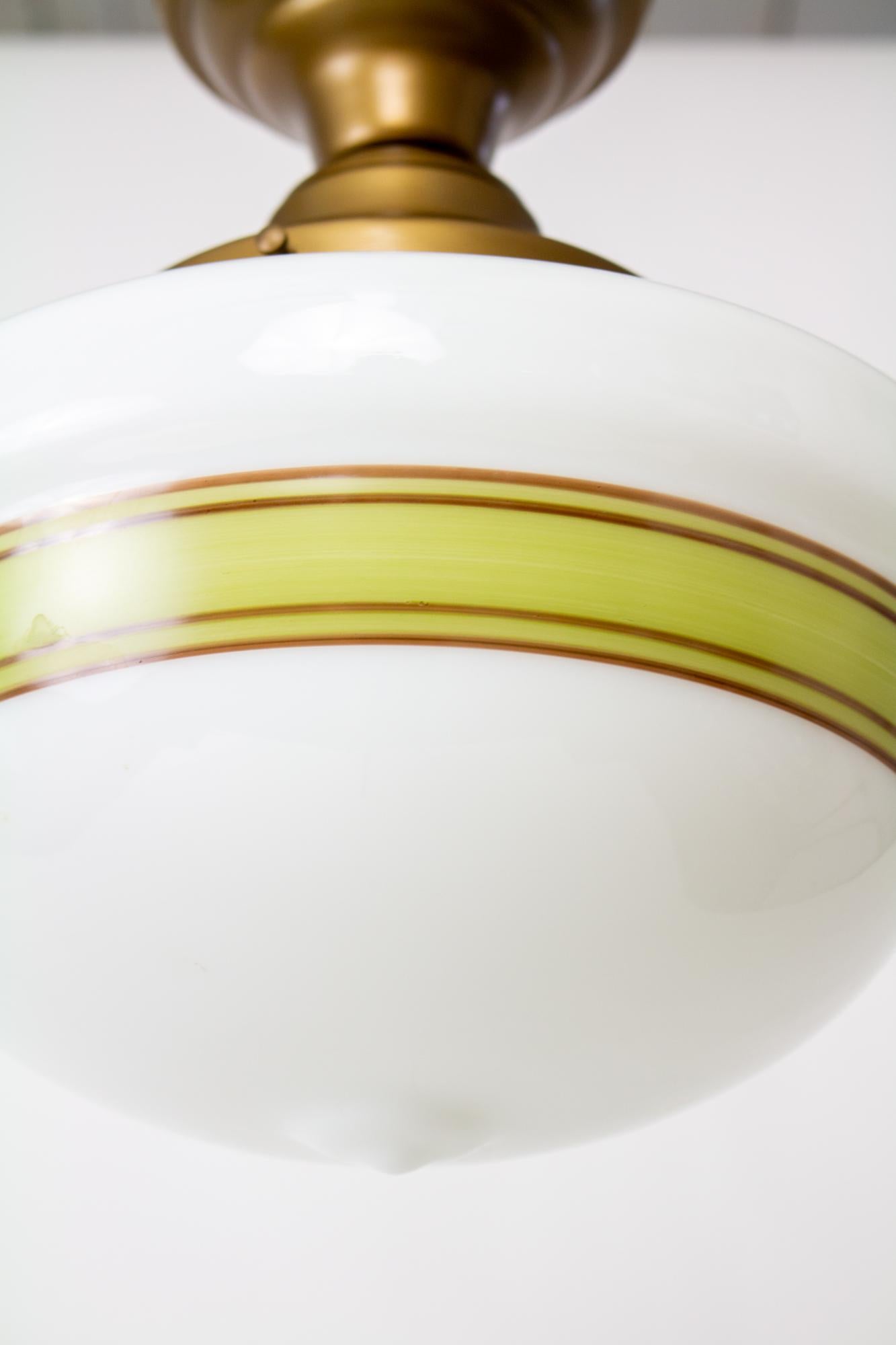 Flush mounted painted schoolhouse fixture. Antique brass finish on new metal fixture. Milk glass in the classic schoolhouse shape with a ring of avocado green and brown stripes. Glass is reproduction, from the early 2000’s. In new condition. US 120V