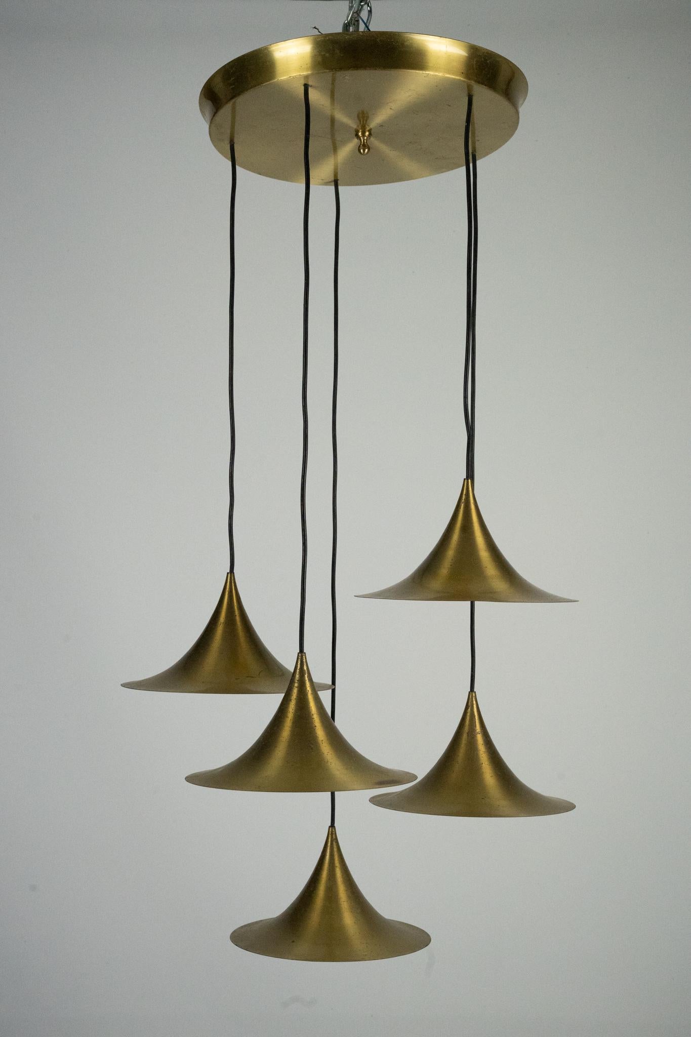 A refined example of Danish midcentury design, this Fog & Morup brass pendant from the 1960s features five elegant conical shades suspended at varying heights from a circular ceiling canopy. The lustrous brass finish and sculptural forms create a