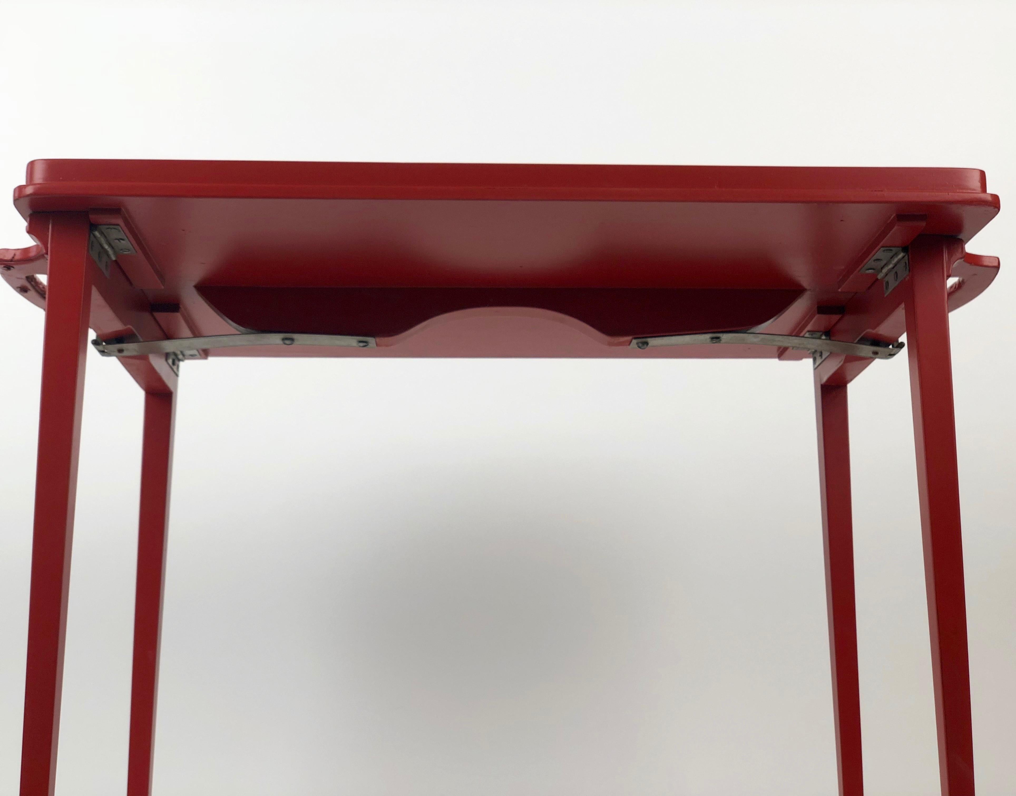 A folding, wooden tray table in burnt orange colour, from the 1930's. The tray uses a simple mechanical mechanism composed of a 
Leaf spring and pin lock. By just pushing the thumb levers it is easy to release the legs and convert the tray to a