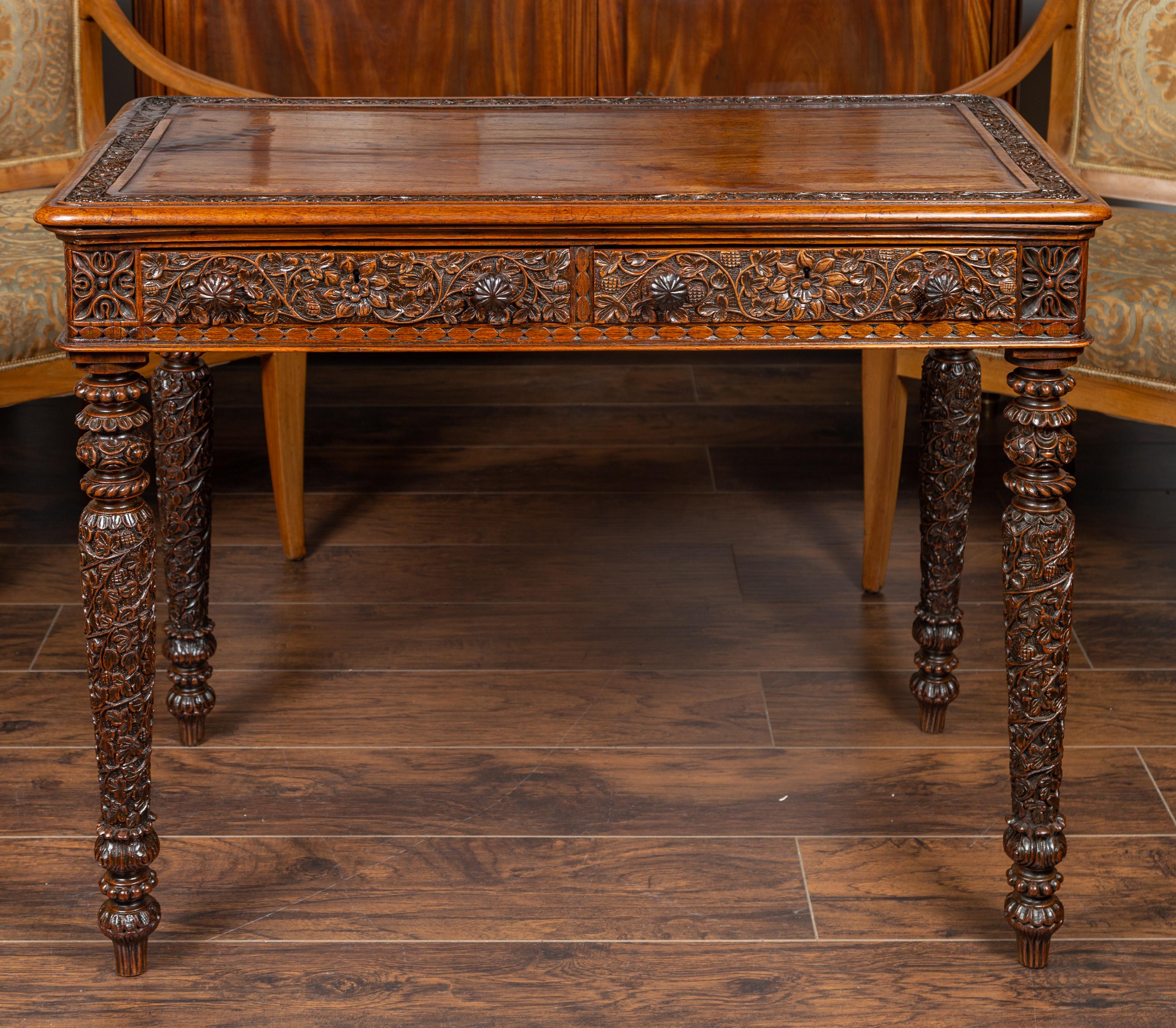 An Anglo-Indian oak table from the early 20th century, with two drawers and richly carved foliage decor. Born at the turn of the century, this exquisite oak table features a rectangular top with rounded corners and delicately carved frame, sitting