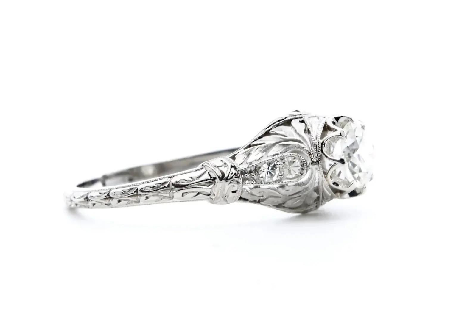 A hand engraved foliate motif Art Deco period diamond engagement ring in platinum. Centered by a 0.70 carat old European cut diamond of H color and VS2 clarity. The hand crafted mounting features leaf motif filigree work complemented by beautiful