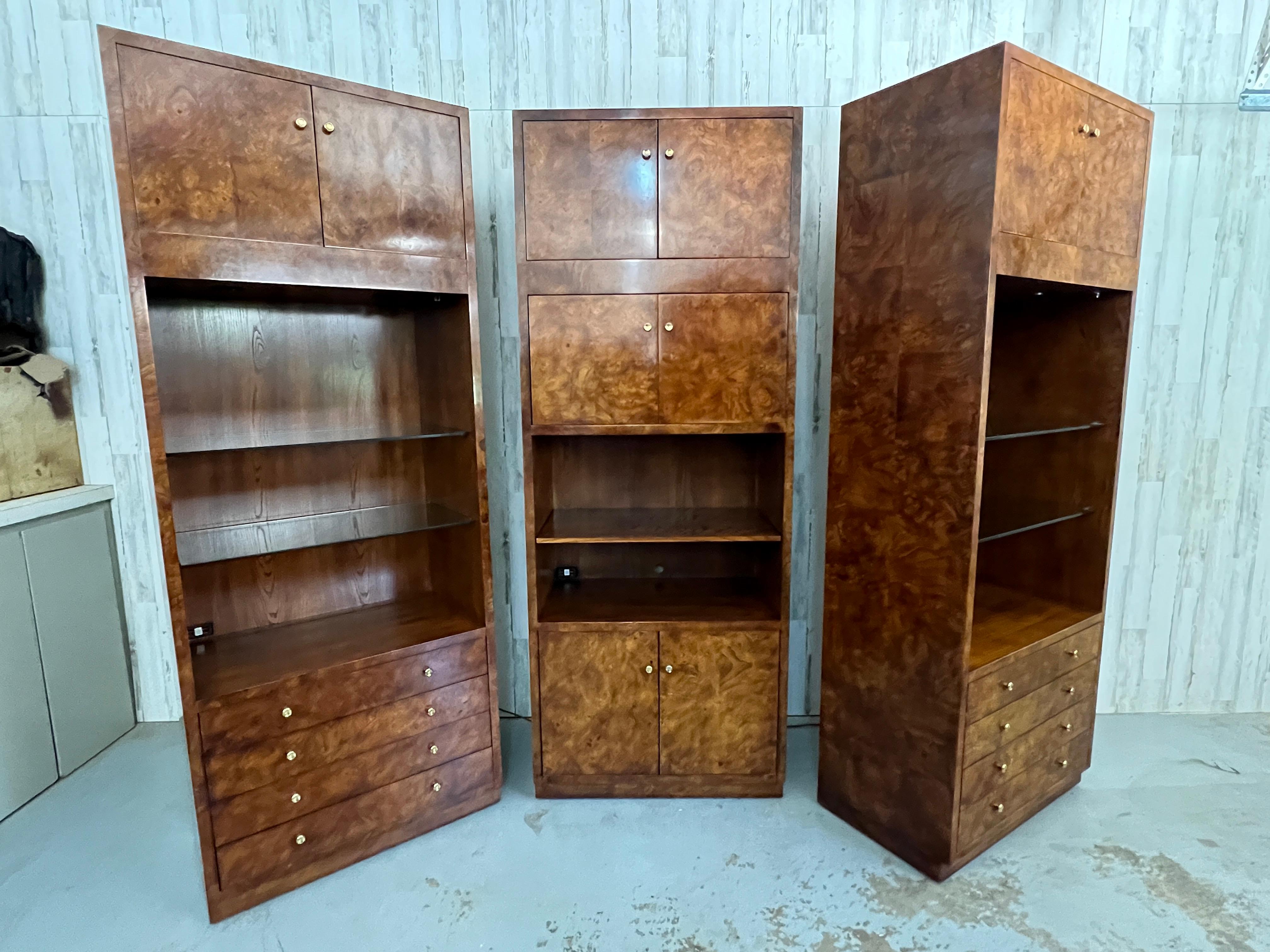 Set of three cabinets with elm burl wood veneer. Recessed lighting in two of the cabinets to illuminate the adjustable glass shelving. Built in electrical outlets for media devices. Brass hardware is a stunning accent to the burl wood.
 Cabinets