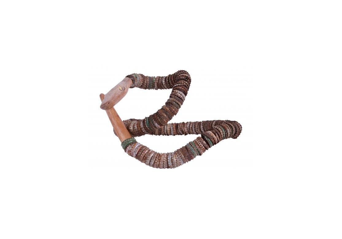 Eine skurrile und überraschend dekorative Schlange aus Dutzenden von bunten Flaschendeckeln, durchbohrt und aufgereiht auf einem starken Metalldraht, der sich zu vielen Figuren drehen und wenden lässt. Der Kopf und der Schwanz sind aus hellem Holz