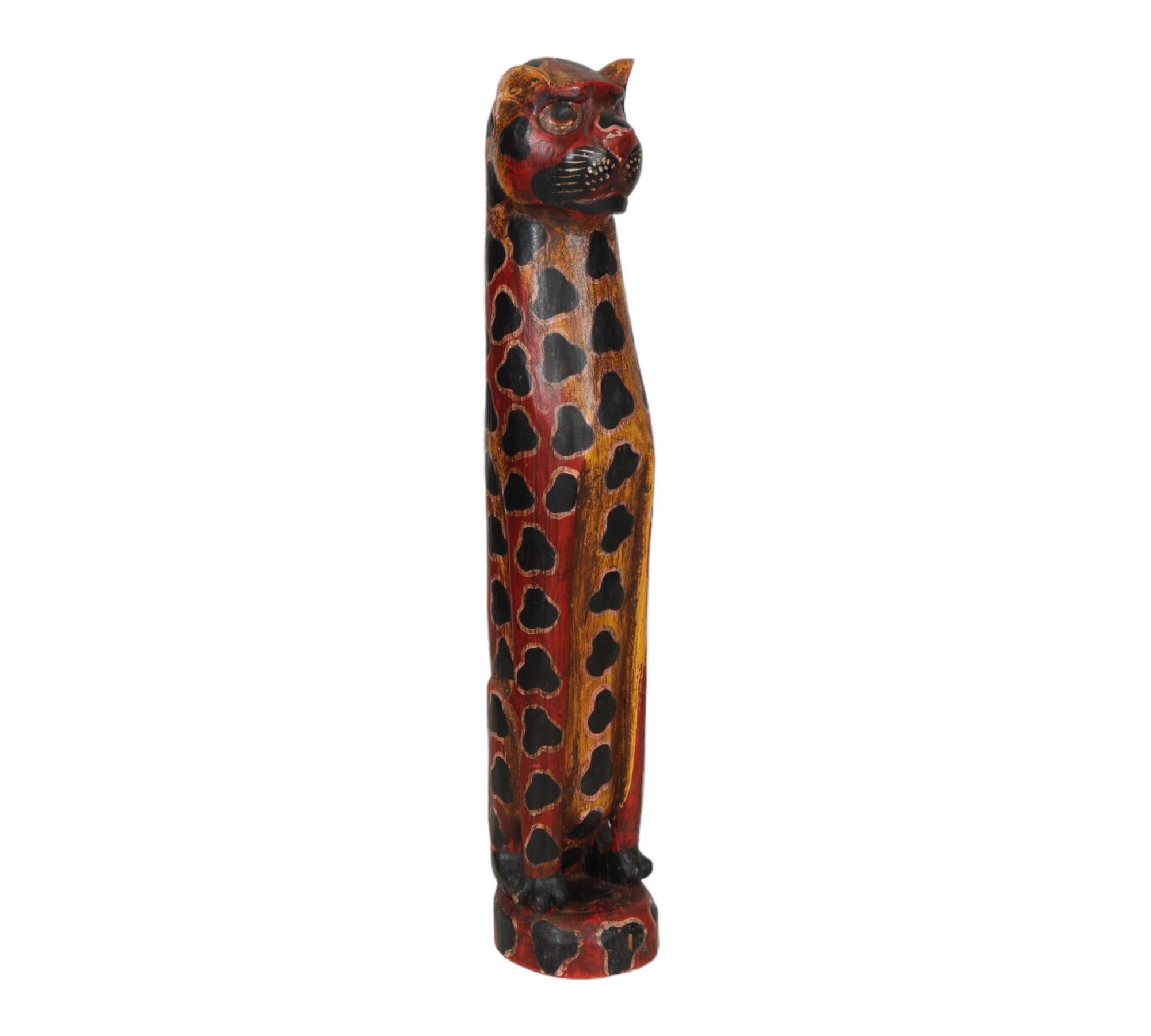 A tall hand carved wooden statue of a seated leopard on a round base. Hand painted throughout in red and yellow with black spots edged in pink. Carved with graceful curves and details defining the features of the face, paws and tail.
