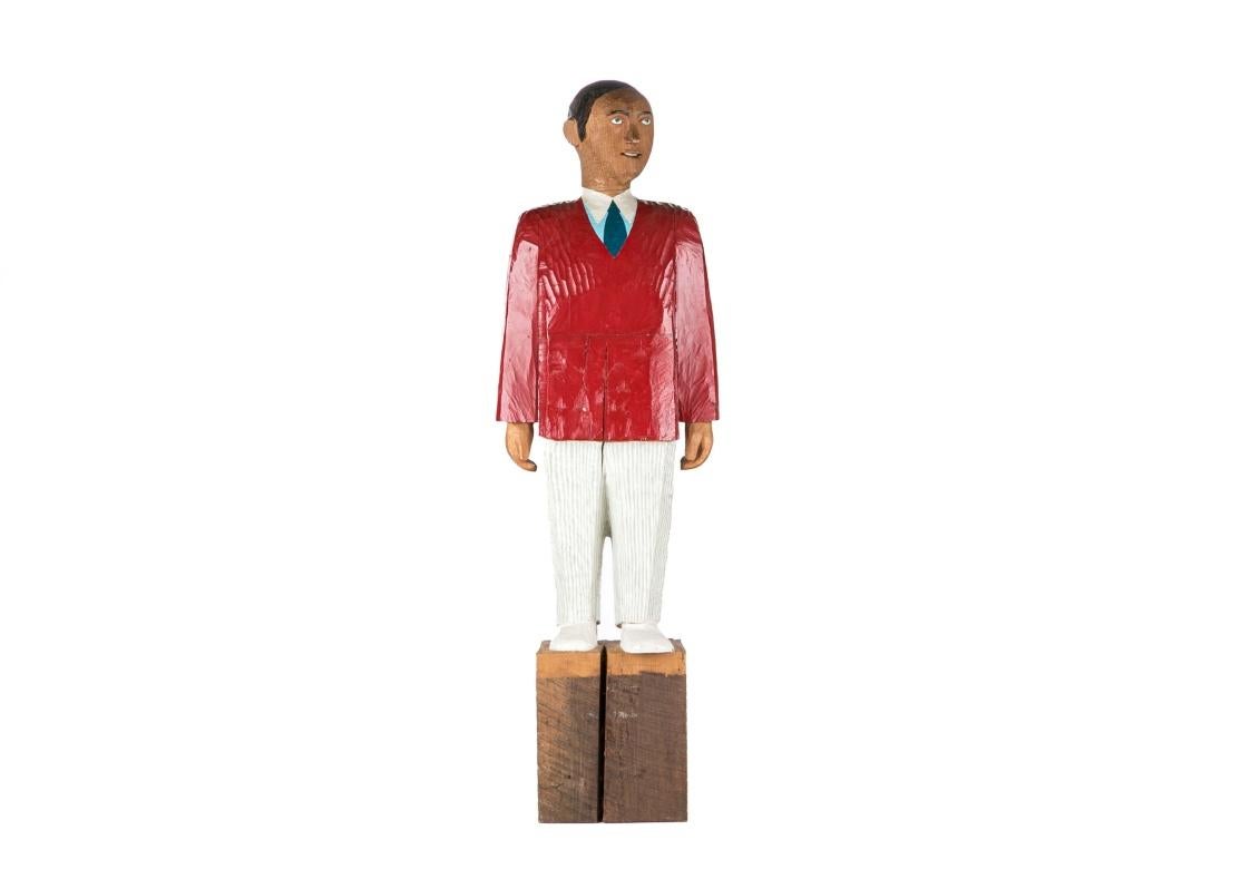 Il s'agit d'une sculpture en bois d'art populaire vintage représentant un homme. La sculpture est peinte et représente un homme vêtu d'une veste rouge, d'un pantalon à rayures blanches et d'une cravate bleue ; elle est surmontée d'un socle.
Incisé