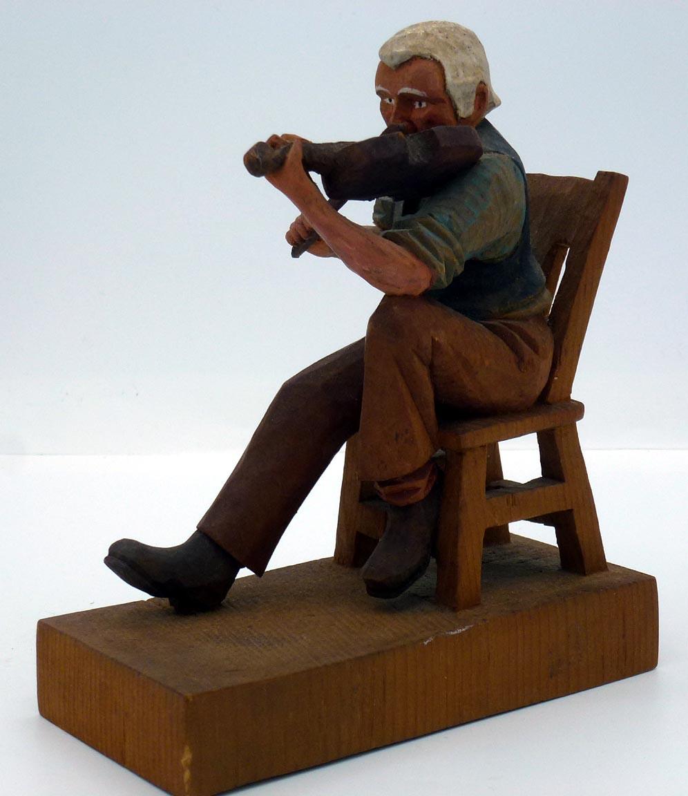 This is a fiddle player with a long mustache, and is seated with one foot propped on his chair. The whole carving, including the chair and base is made from a single piece of wood. It is 5 5/8