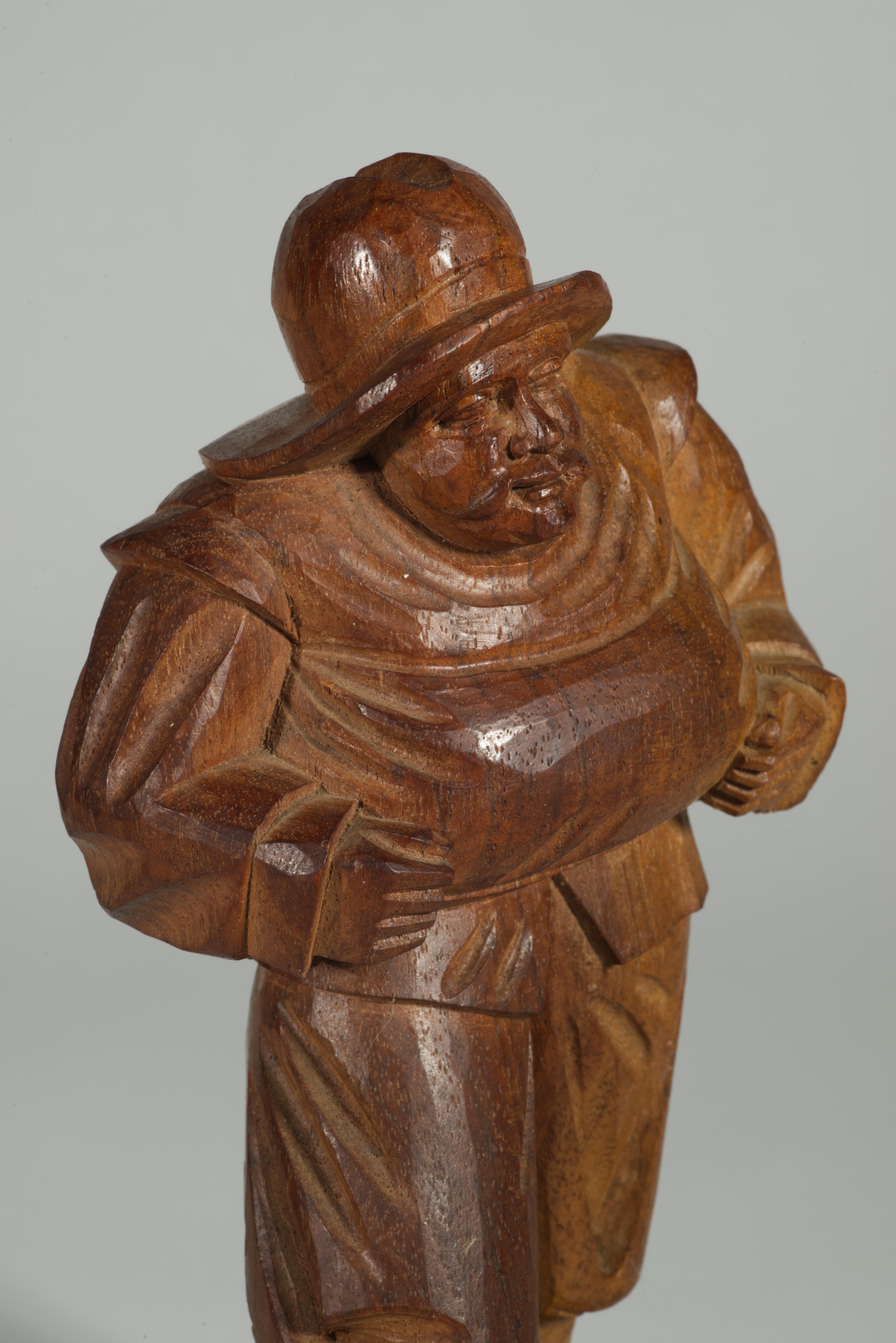Escultura folclórica primitiva de madera tallada a mano que representa a un hombre que camina con ropas amplias y holgadas y sombrero, llevando un fardo en las manos. El tallado muy detallado resalta el veteado natural de la madera y otras