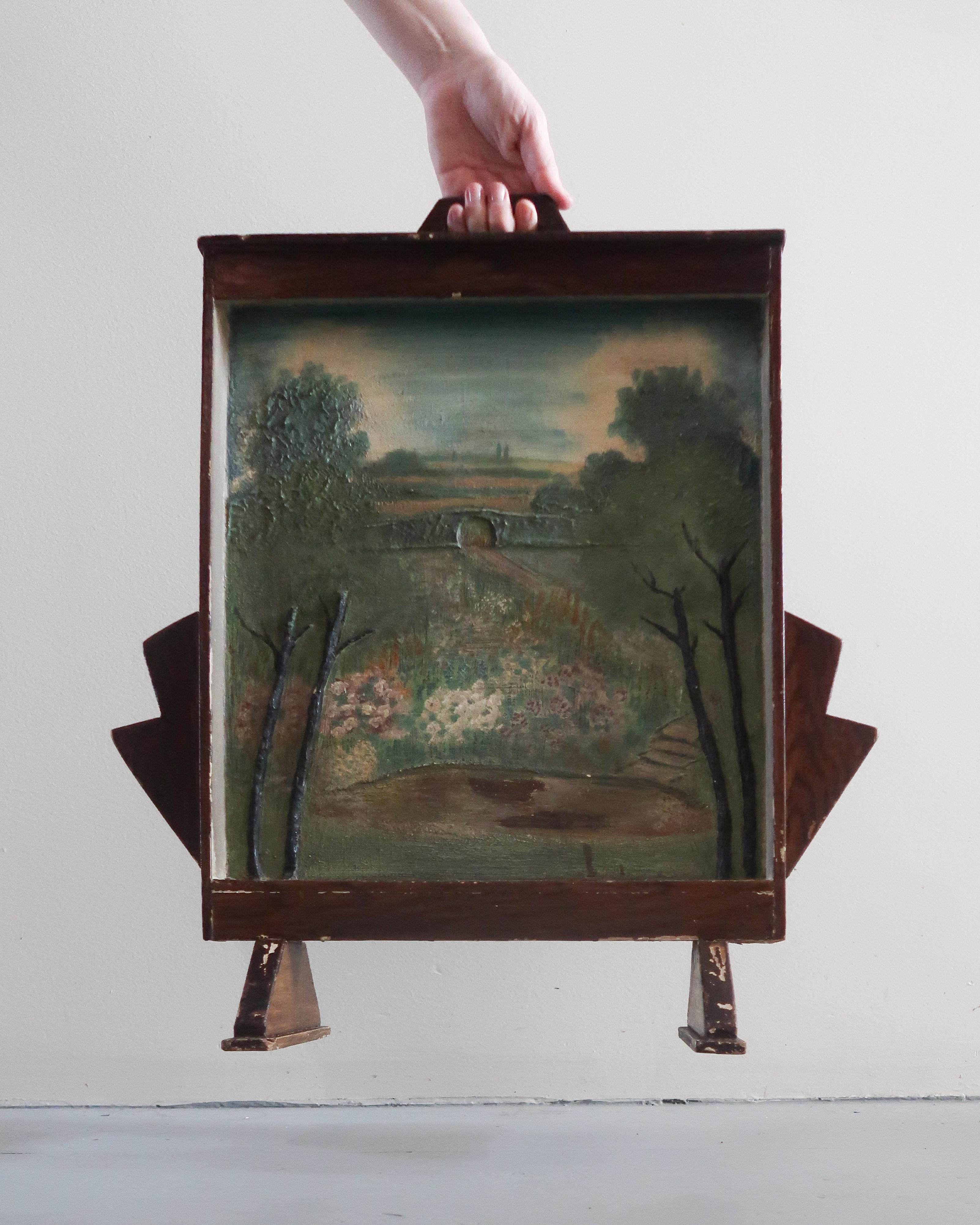 Wood fireplace screen featuring a hand-painted folk art landscape scene on canvas. The frame is solid wood with angular side panels and short feet, giving the piece a sculptural presence. The pastoral scene shows trees, a bridge, and a moody sky