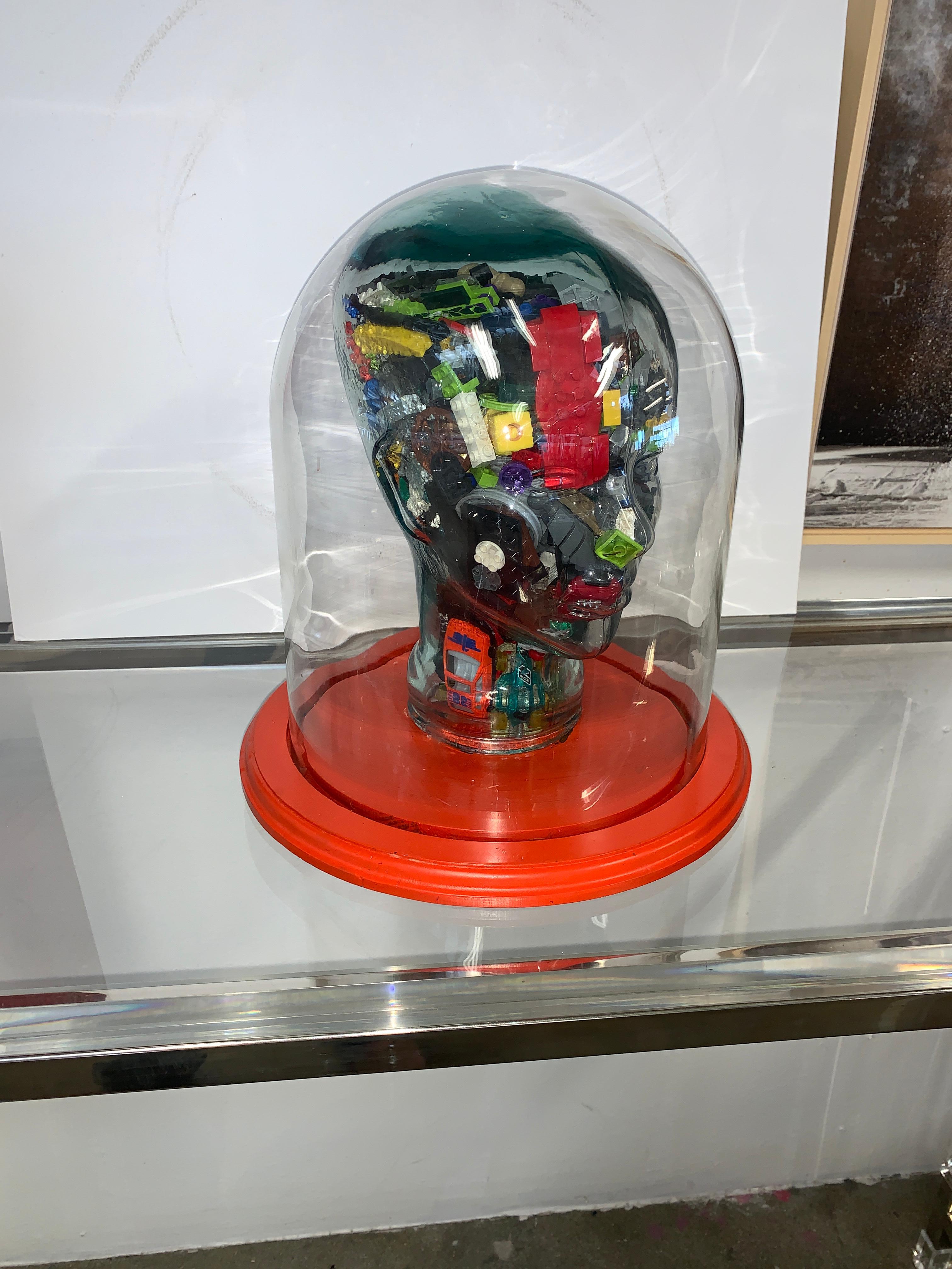 A Folk Art or Outsider Art bust under a glass dome. The glass bust is molded and it is filled with all sorts of found objects, including legos, chess pieces and blocks among other things. Likely late 20th or early 21st century. The base is a shade