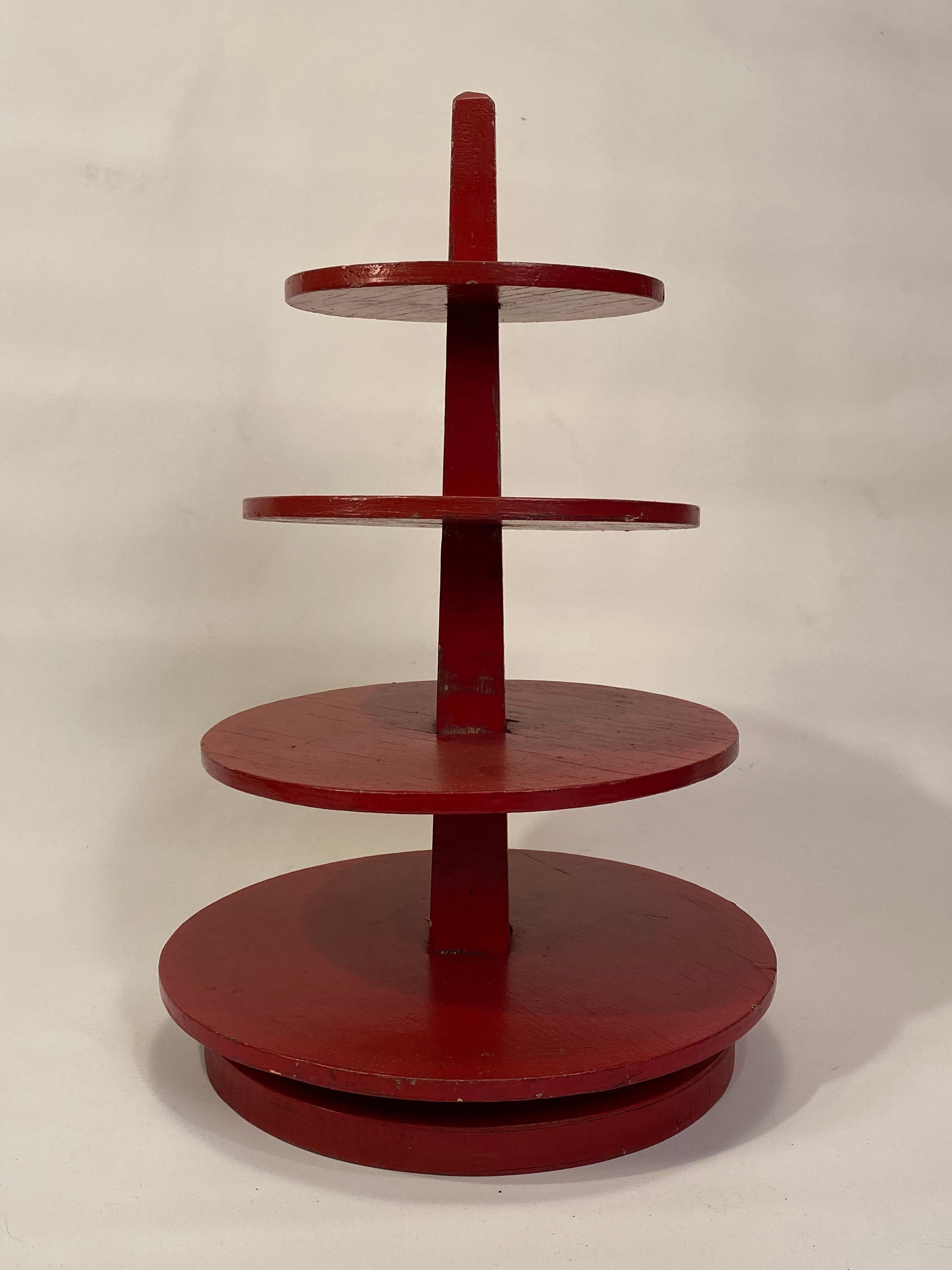 Charming red enamel painted tiered plywood display stand. A wonderful way to display your collectibles and curios. Comprised of four round tiered plywood shelves that rest snuggly around a center post (it does not rotate). There is no glue so one or