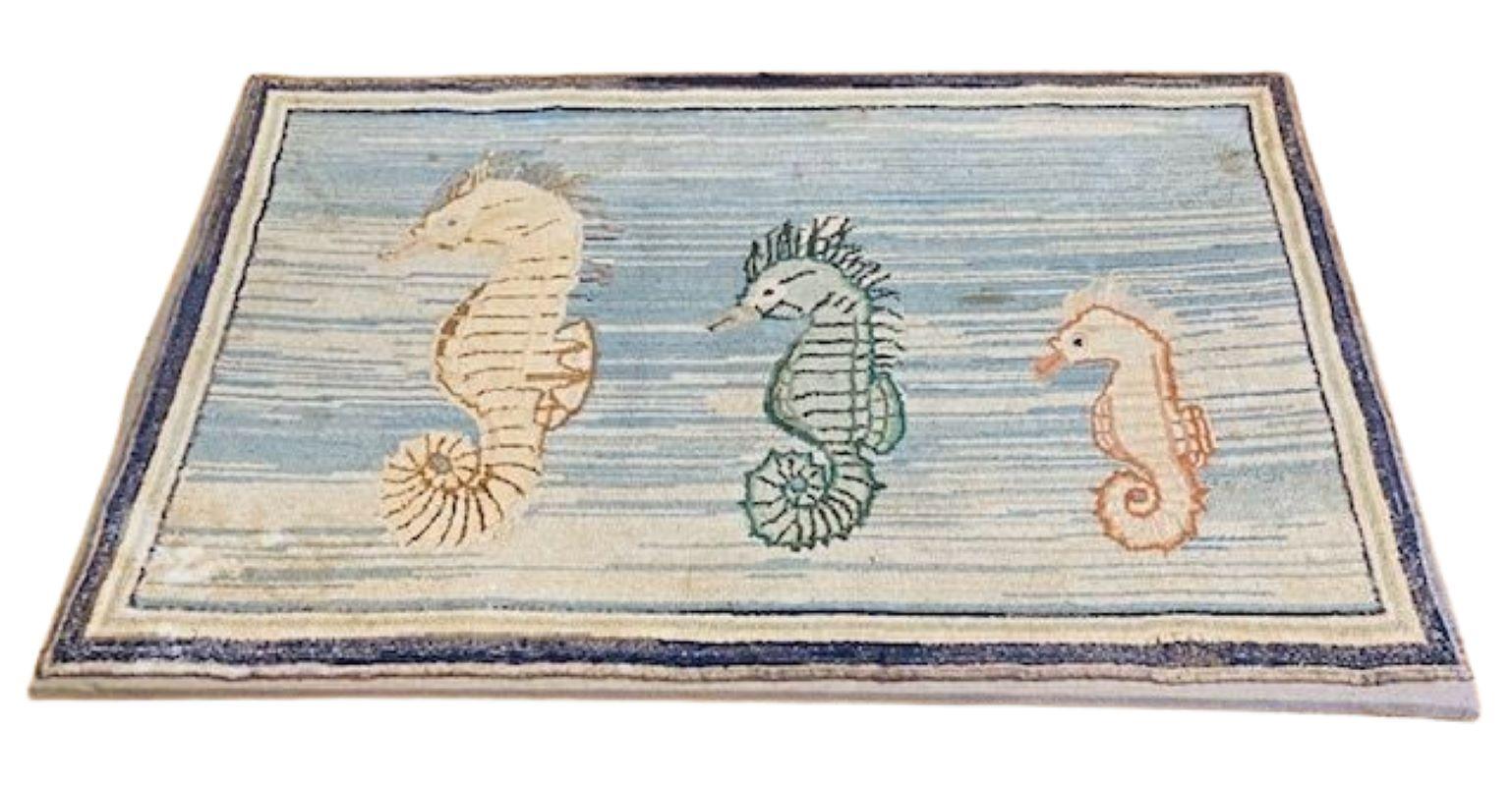 Tapis crocheté Vintage Folk Art Sea Horse, circa 1960, représentant une famille de trois hippocampes de tailles et de couleurs variées nageant sur un fond d'océan bariolé bleu et blanc, monté sur un châssis en bois recouvert de lin pour être