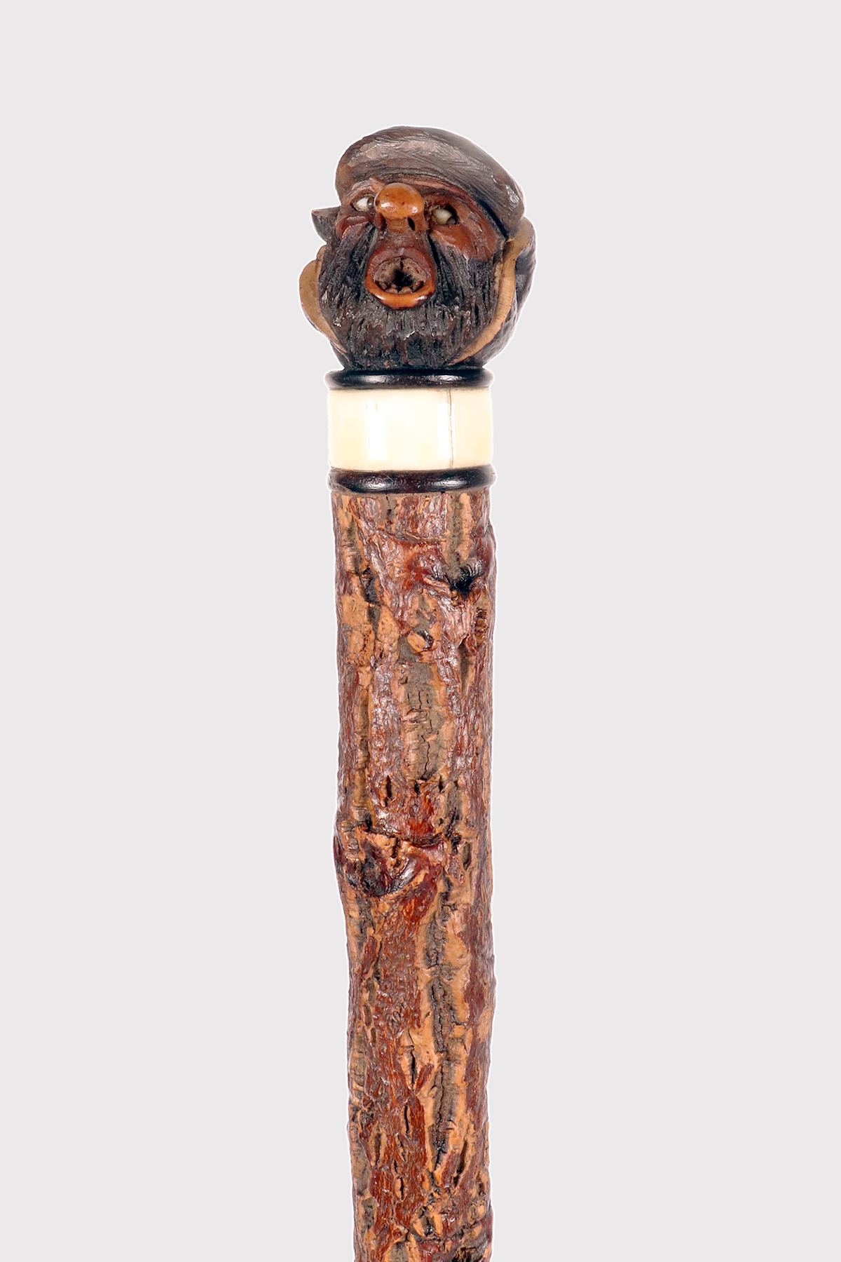 Bastone da passeggio in stile folk: ramo singolo in legno di sughero. Il manico del bastone, un dado corozo intagliato che rappresenta la testa di un uomo grottesco con un viso oblungo con la bocca aperta e gli occhi in pasta di vetro. Anello in