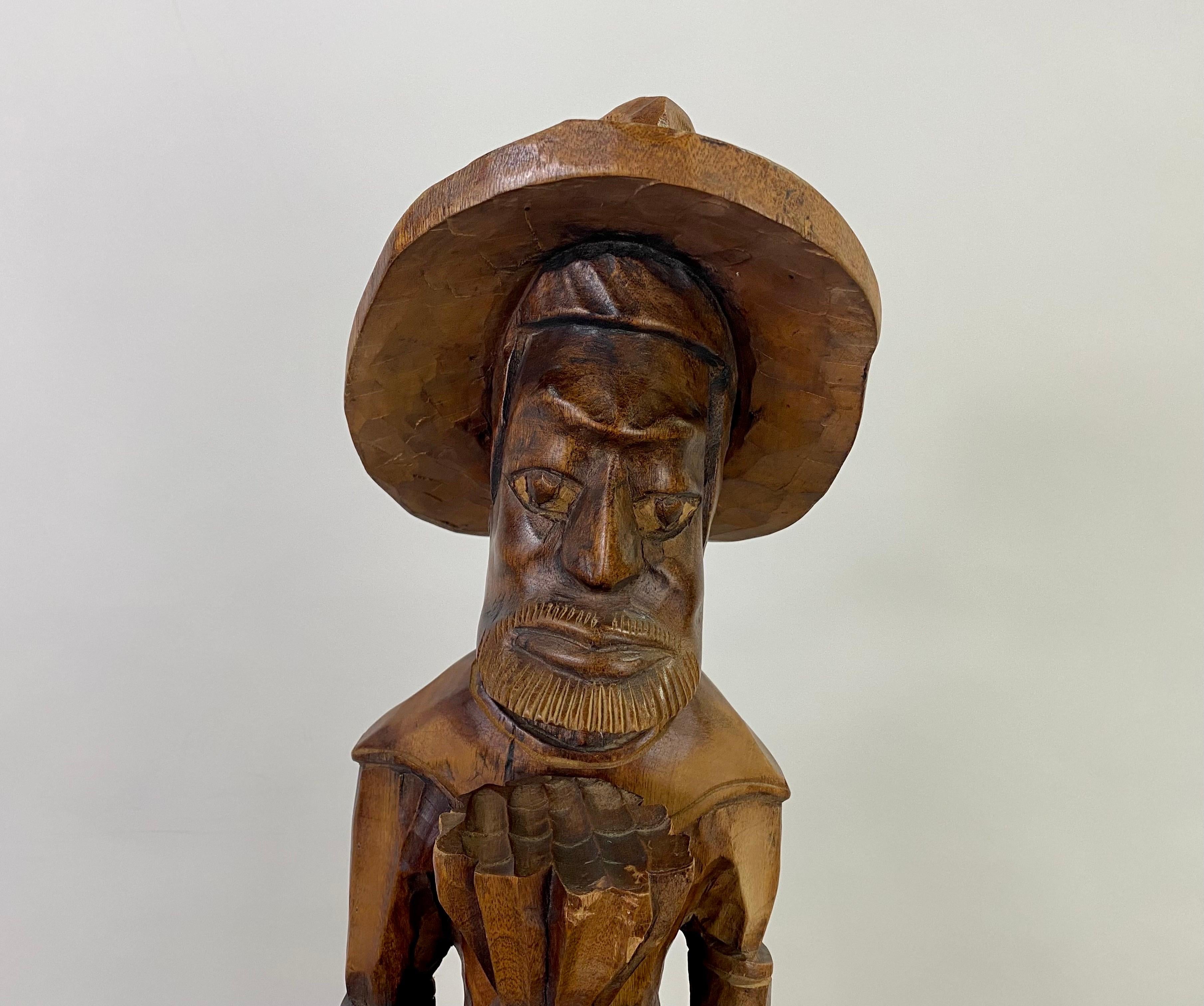 A vintage Mid-Century period Folk art sculpture of a man standing tall.  The sculpture is finely carved of quality wood and depicts a Caribbean farmer man wearing a hat, holding a pineapple with one hand and an umbrella in the other hand. 