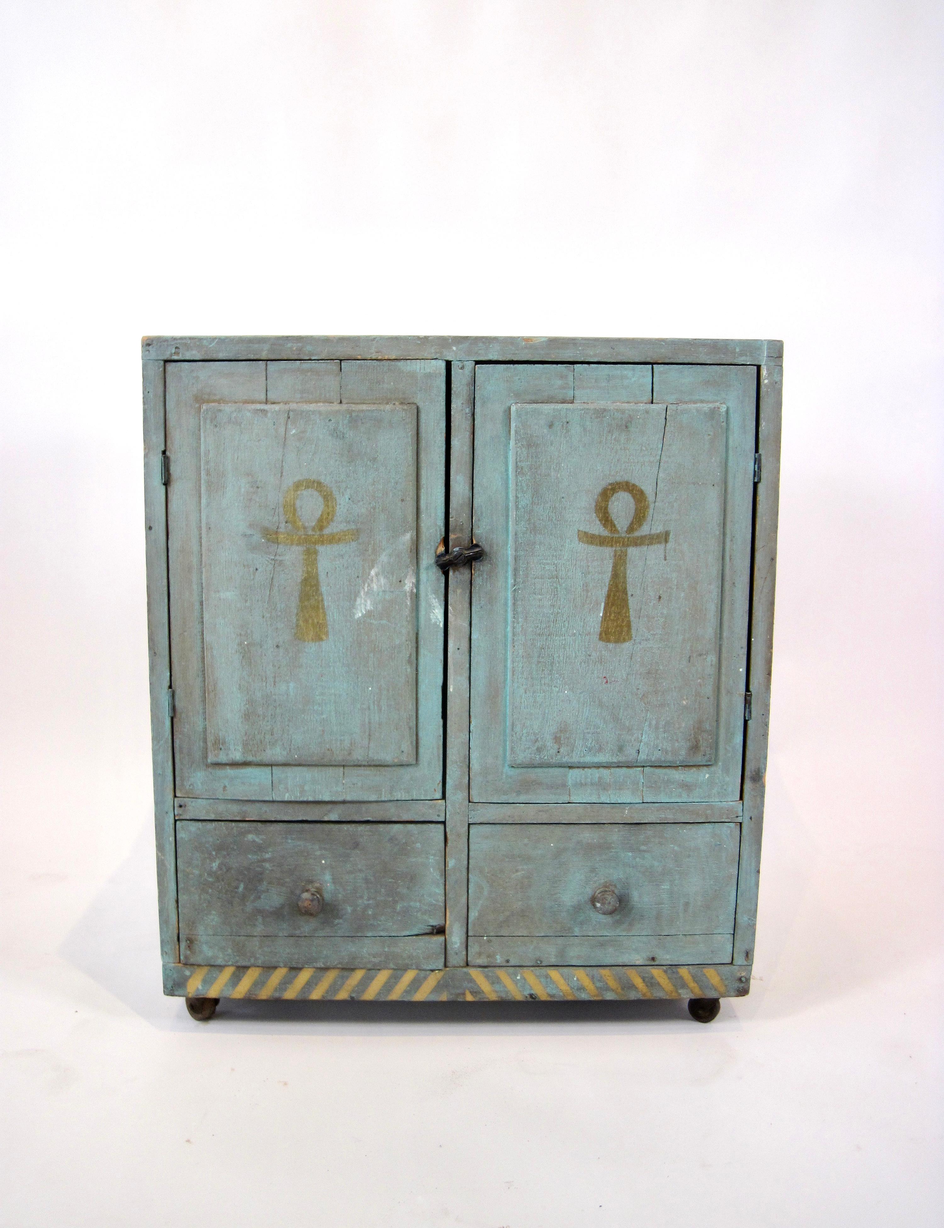 Armadietto in legno di recupero in stile folkloristico e verdastro chiaro con simboli egizi dorati su rotelle. Due ante dell'armadio con simboli ankh e due cassetti sottostanti. All'interno quattro cassetti con dipinti i simboli dell'Occhio di Ra e