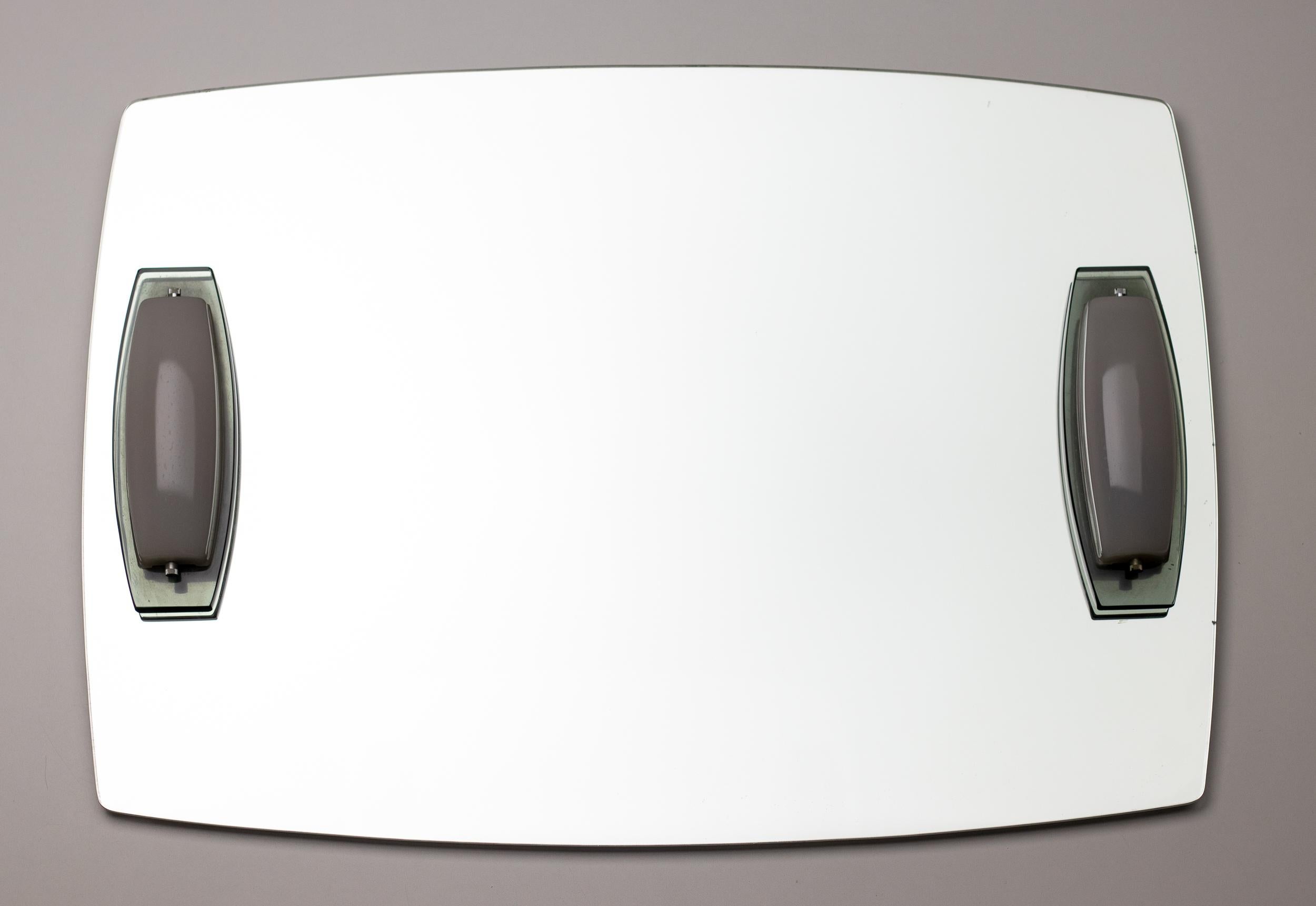 Un superbe miroir avec 2 lumières intégrées de Max Ingrand pour Fontana Arte.
Les lampes sont conçues avec une base biseautée vert émeraude typique de Max Ingrand et un abat-jour en verre de couleur blanche. Elles sont équipées de douilles E14