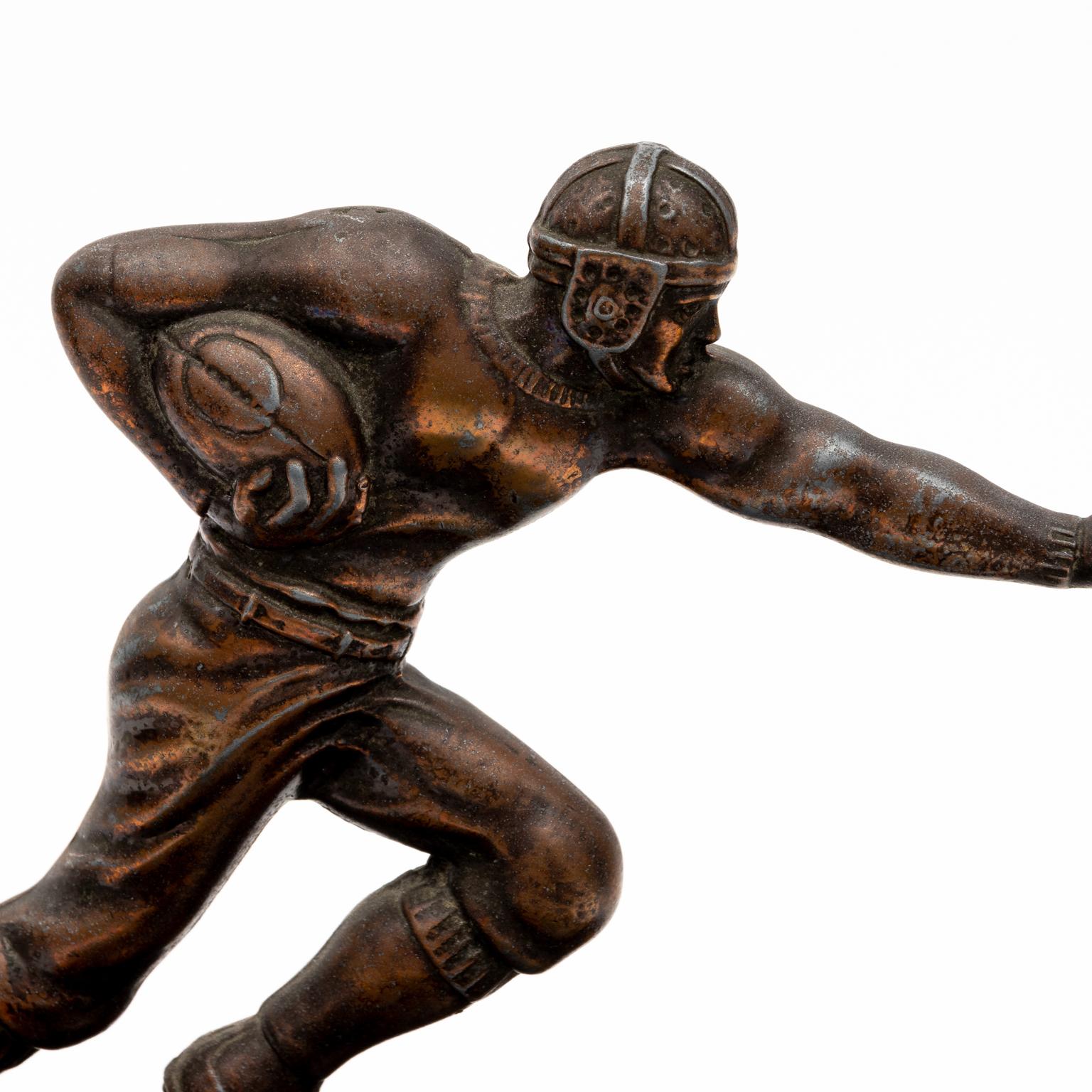 Circa 1920-1930s rare football mascot car radiator cap or hood ornament in cast metal. The piece looks like a pre-Heisman trophy style stance with left arm up and right arm tucked. The football player is wear a vented dog eared helmet with leather