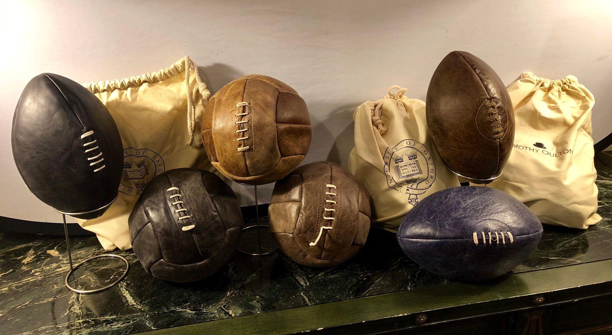 A group of seven footballs and soccer balls by Timothy Oulton. Each ball comes with a 'oxford university' canvas bag, and a pin to inflate. All are worn as is the intended and desired look and design. Perfect to decorate an office, poolroom,