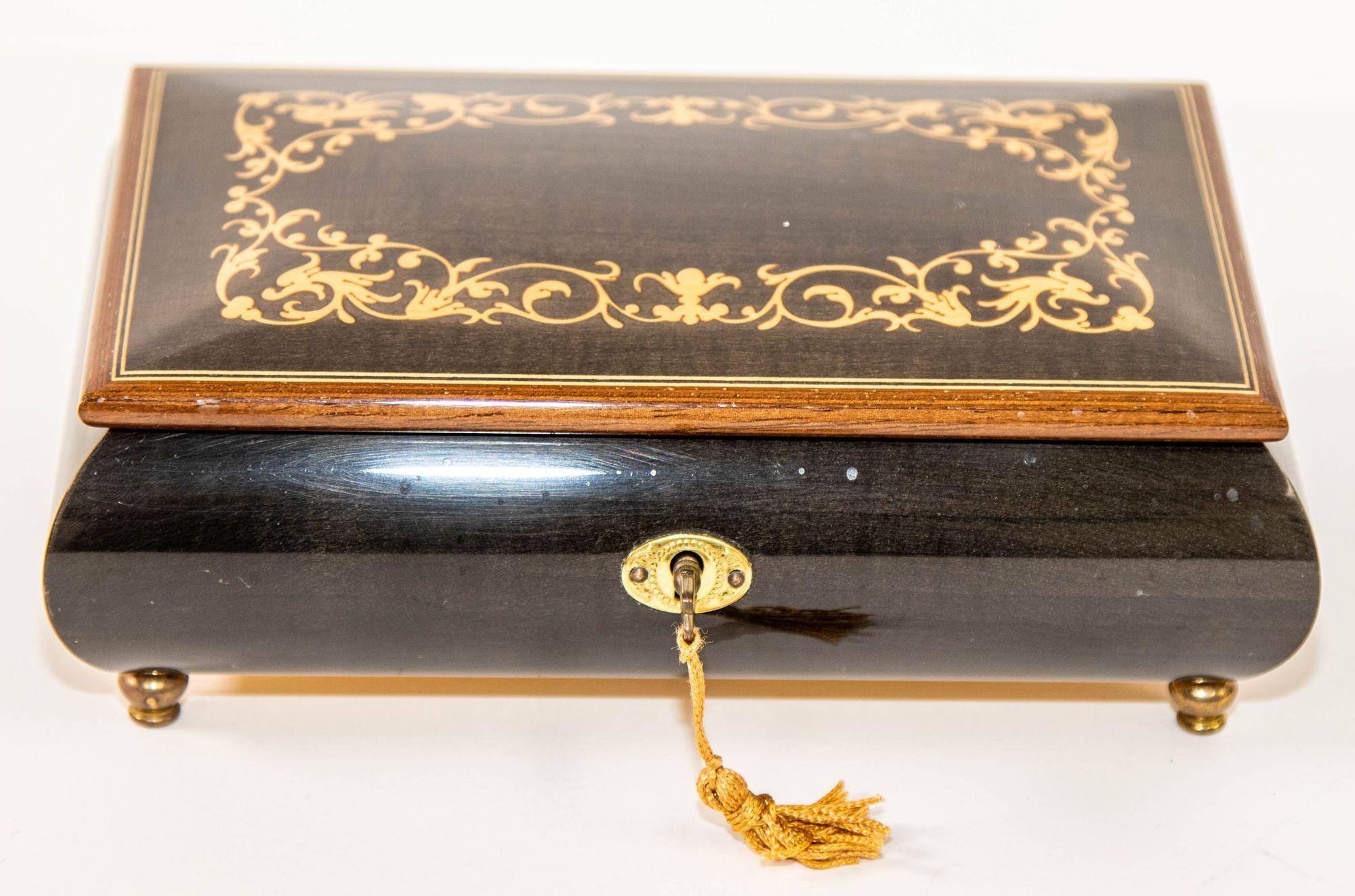 Elegante caja de música de madera con pie, de color azul oscuro, con la parte superior finamente pintada a mano y decorada con incrustaciones de follajes florales dorados.
Caja de música artesanal con pie de madera de thuya pintada a mano en la