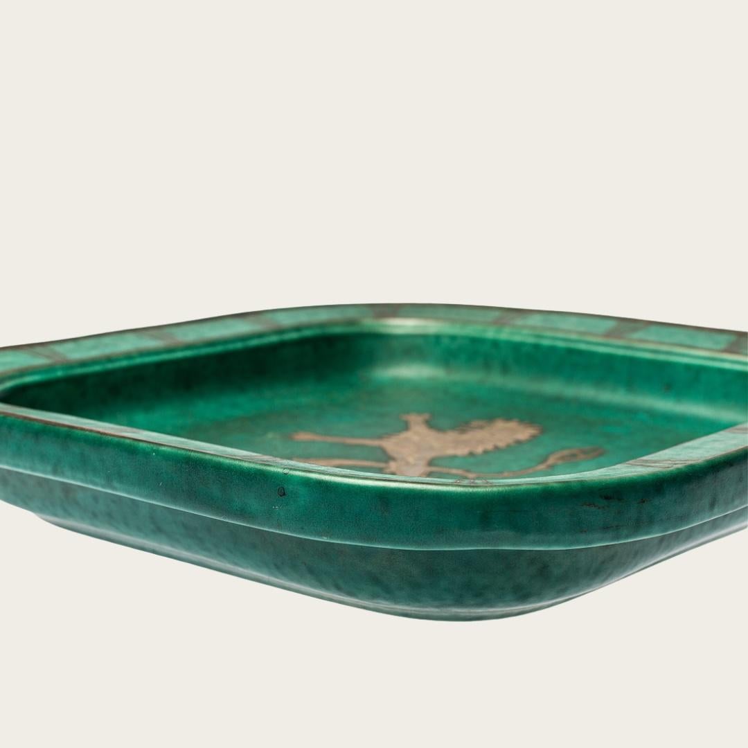 1972, Sweden. Footed dish part of Wilhelm Kåge’s Argenta stoneware collection made for the Gustavsberg Factory. It features a speckled turquoise glaze, slightly darker than typical, with silver leaf accents depicting a lion at the center. The dish