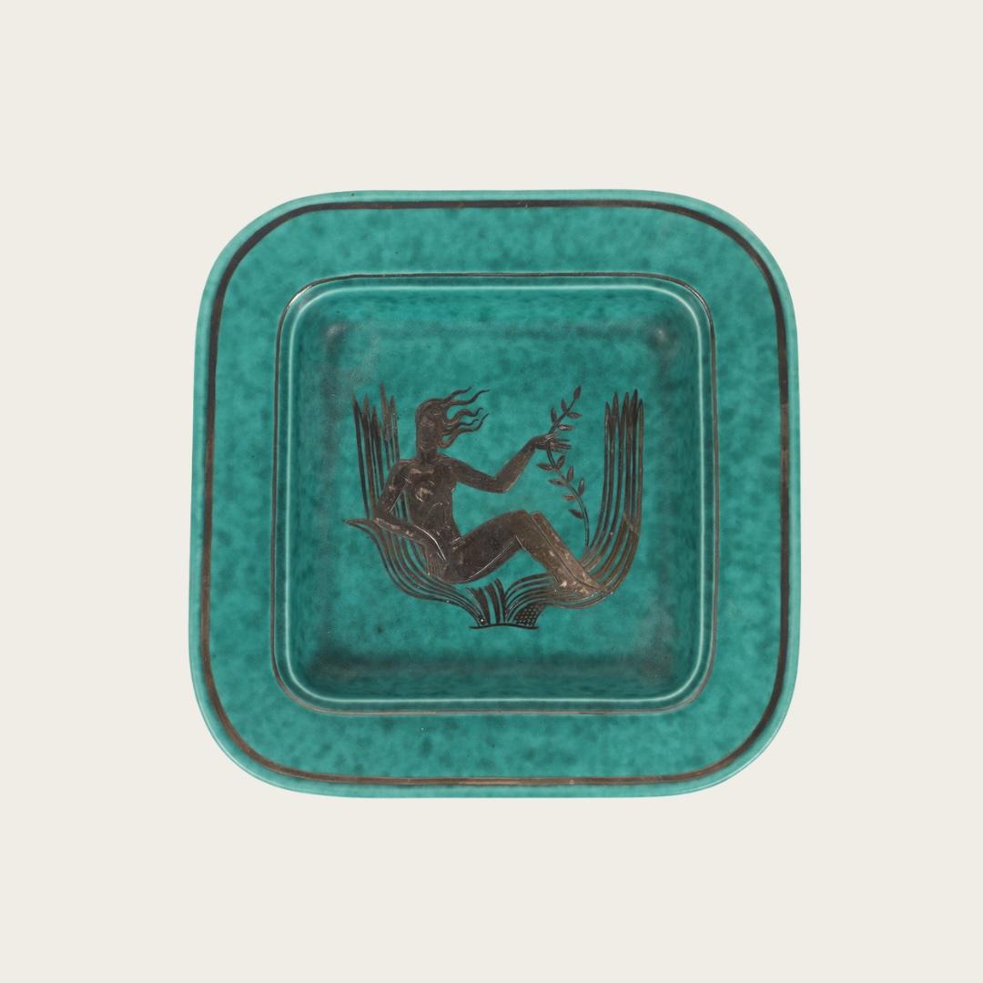 1972, Sweden. This footed dish is a striking example of Wilhelm Kåge’s Argenta collection for Gustavsberg Factory. Crafted in stoneware, it features a speckled turquoise glaze and silver leaf detailing that highlights an Art Deco mermaid at its