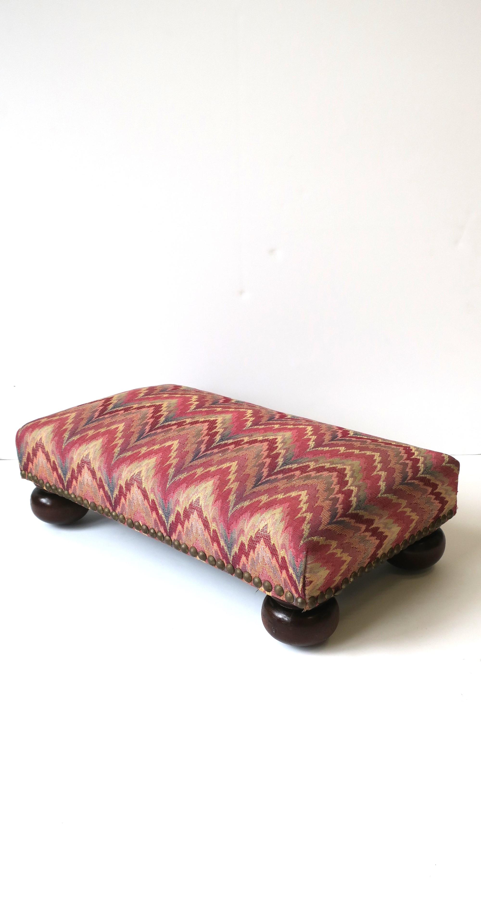 A footstool with rich brown bun feet, upholstered in (possibly) a Schumacher flame-stitch fabric, with brass nail-head design around, circa mid-20th century. Great as a footstool or low bench seat. Piece can also display books, hold a cocktail,