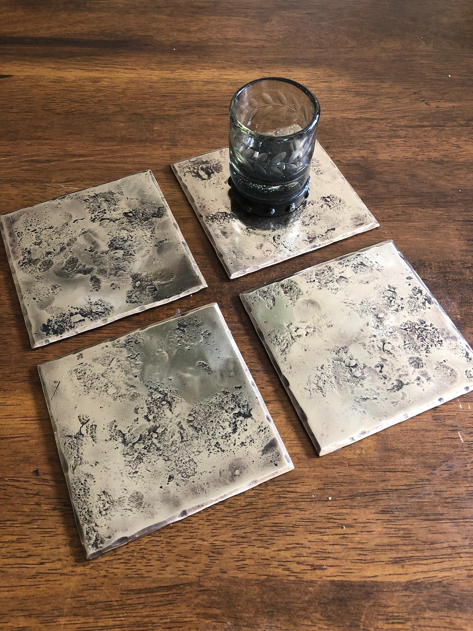 A handcrafted, square coaster made from bronze that has been forge textured over a rough steel anvil face to provide texture. The edges are hammered and beveled. The bronze is burnished to accentuate the forged details. A glossy clear coat enhances