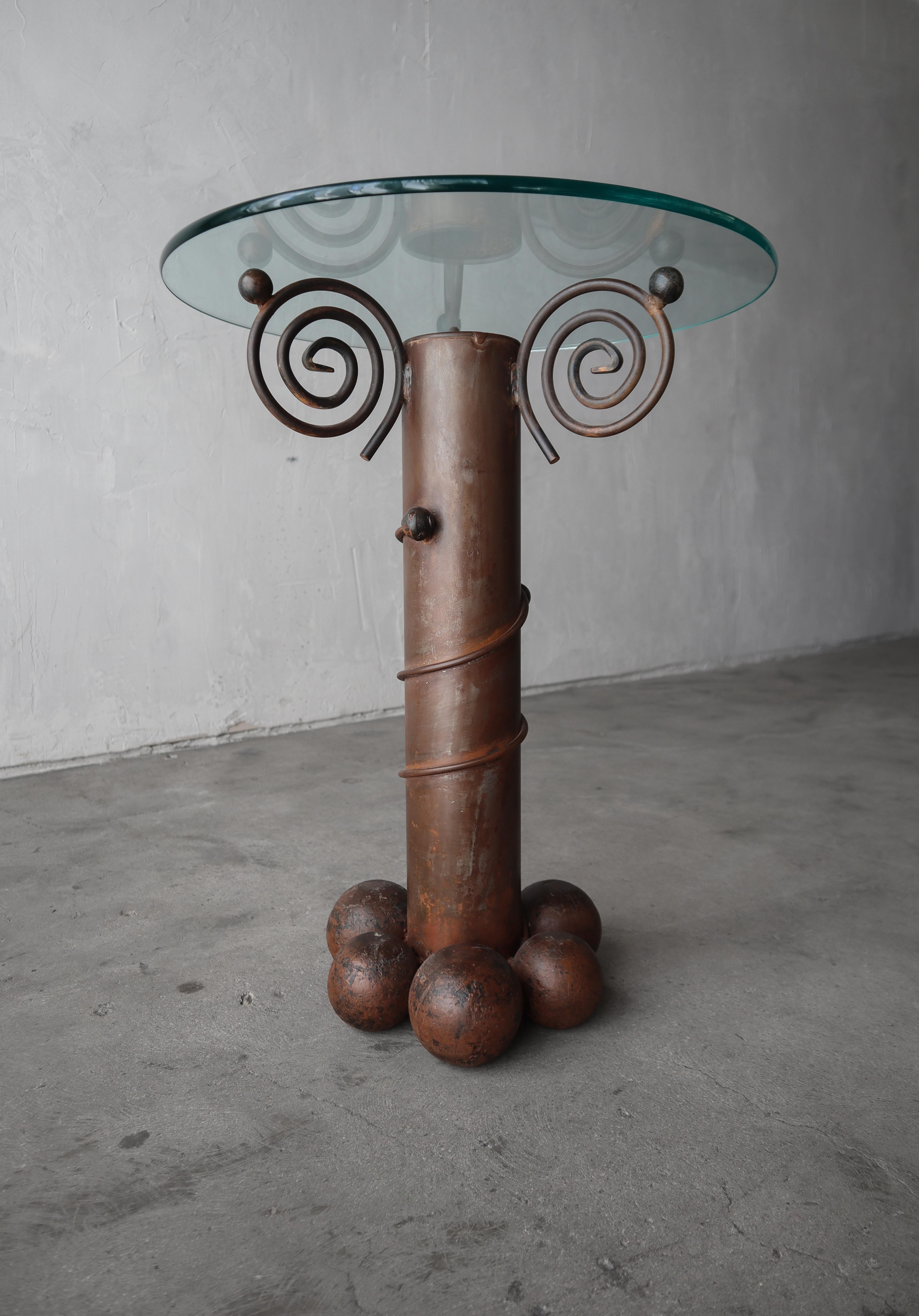 Unique, handmade side table by Carlos De Anda and made is Mexico. The table base is constructed of forged iron with center column design supported by large, solid iron ball feet and scroll details that support a glass top. Very artistic piece. All