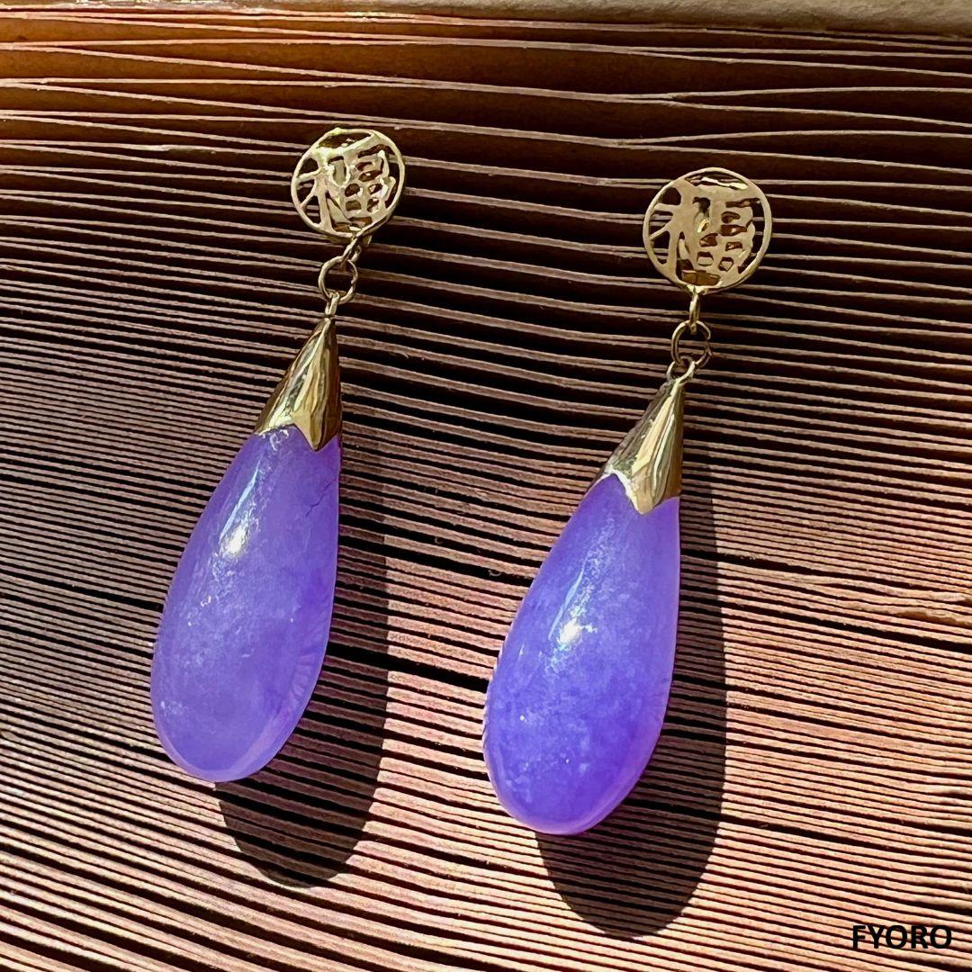 Les boucles d'oreilles Fu Fuku Fortune en jade violet sont dotées d'un fermoir portant le symbole de la fortune et d'un design classique en forme de goutte d'eau. Ces amulettes sont des symboles qui protègent et soutiennent vos objectifs du début à
