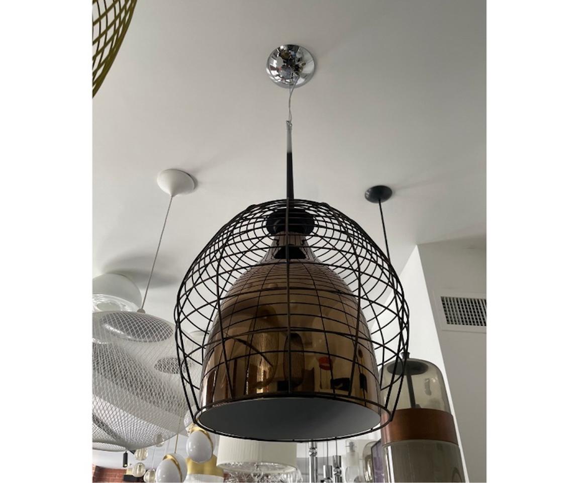 Il Ciondolo Cage è un design vintage con una gabbia irregolare in acciaio circondata da un diffusore in vetro soffiato. Il Ciondolo Cage è caratterizzato da una gabbia esterna in nero opaco e proietta un intenso fascio di luce che attira