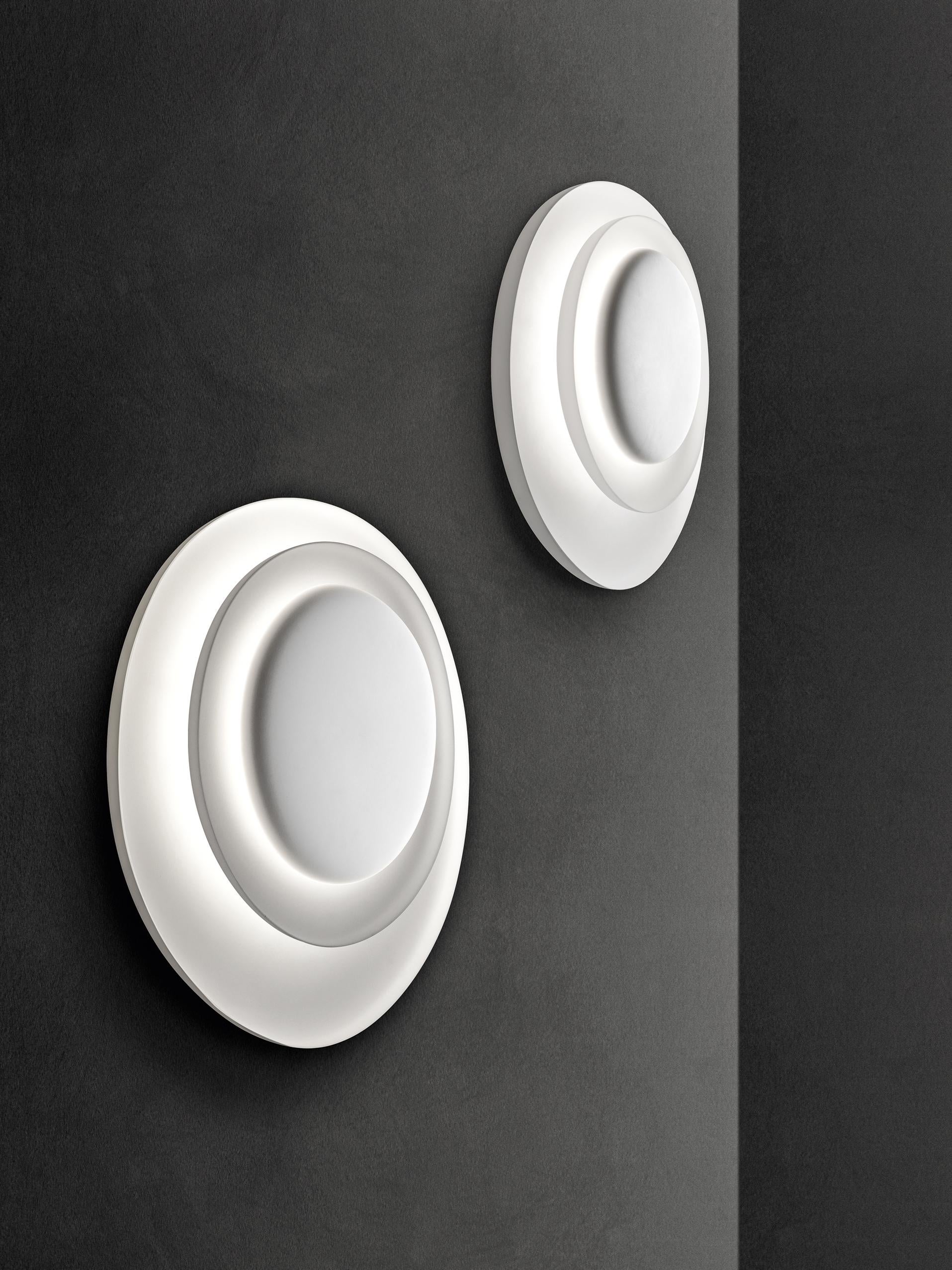 Wall lamp with reflected and diffused light. Consisting of three matte white injection molded polycarbonate egg-shaped plates and fitted together asymmetrically. The largest plate works as the wall mounting plate, and the two central plates conceal