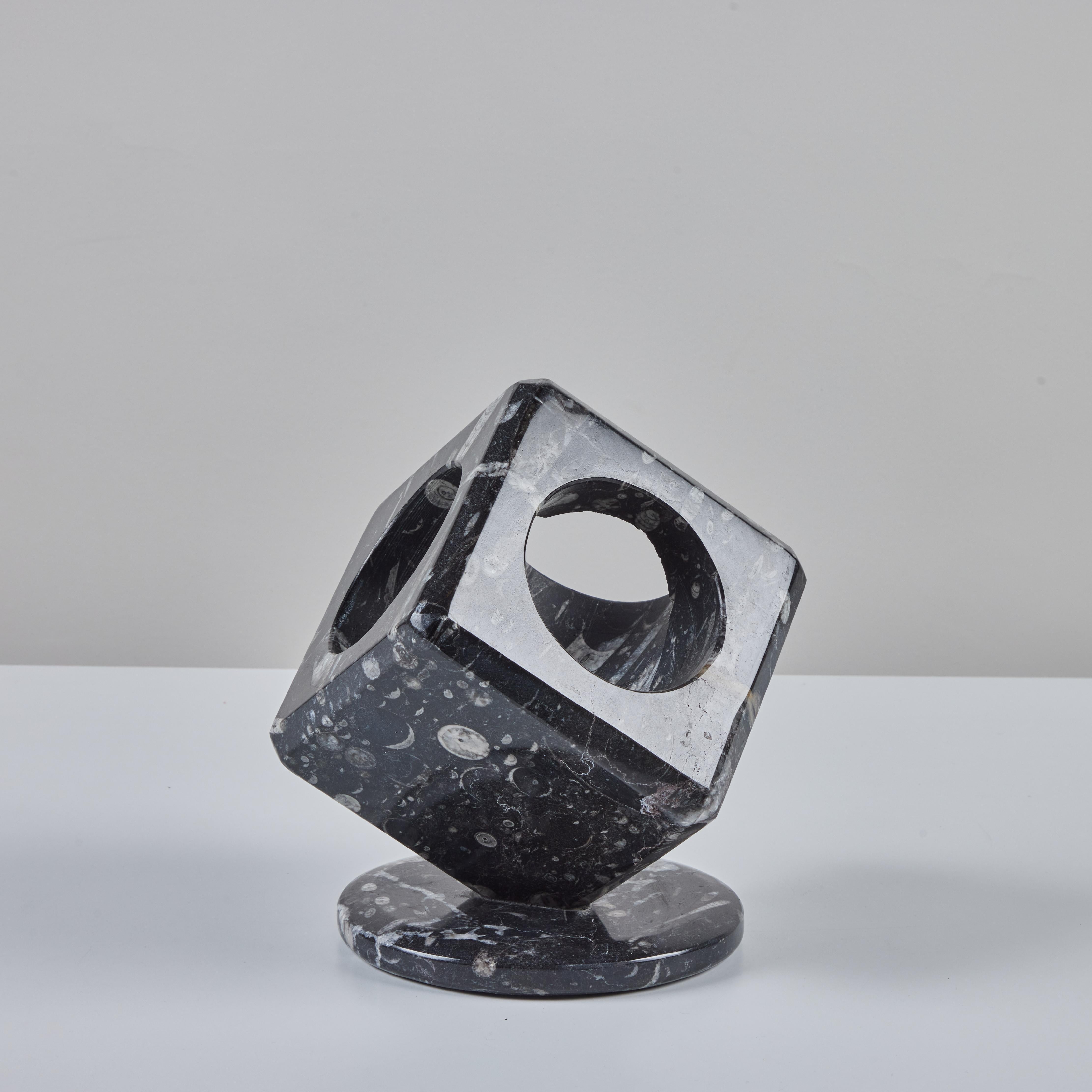 Black marble sculpture set on a round marble base. The sculpture features a polished exterior stone with cream veining and fossils throughout. Three sides of the cubic sculpture have a circular cutouts and hollowed out interior, which showcases a