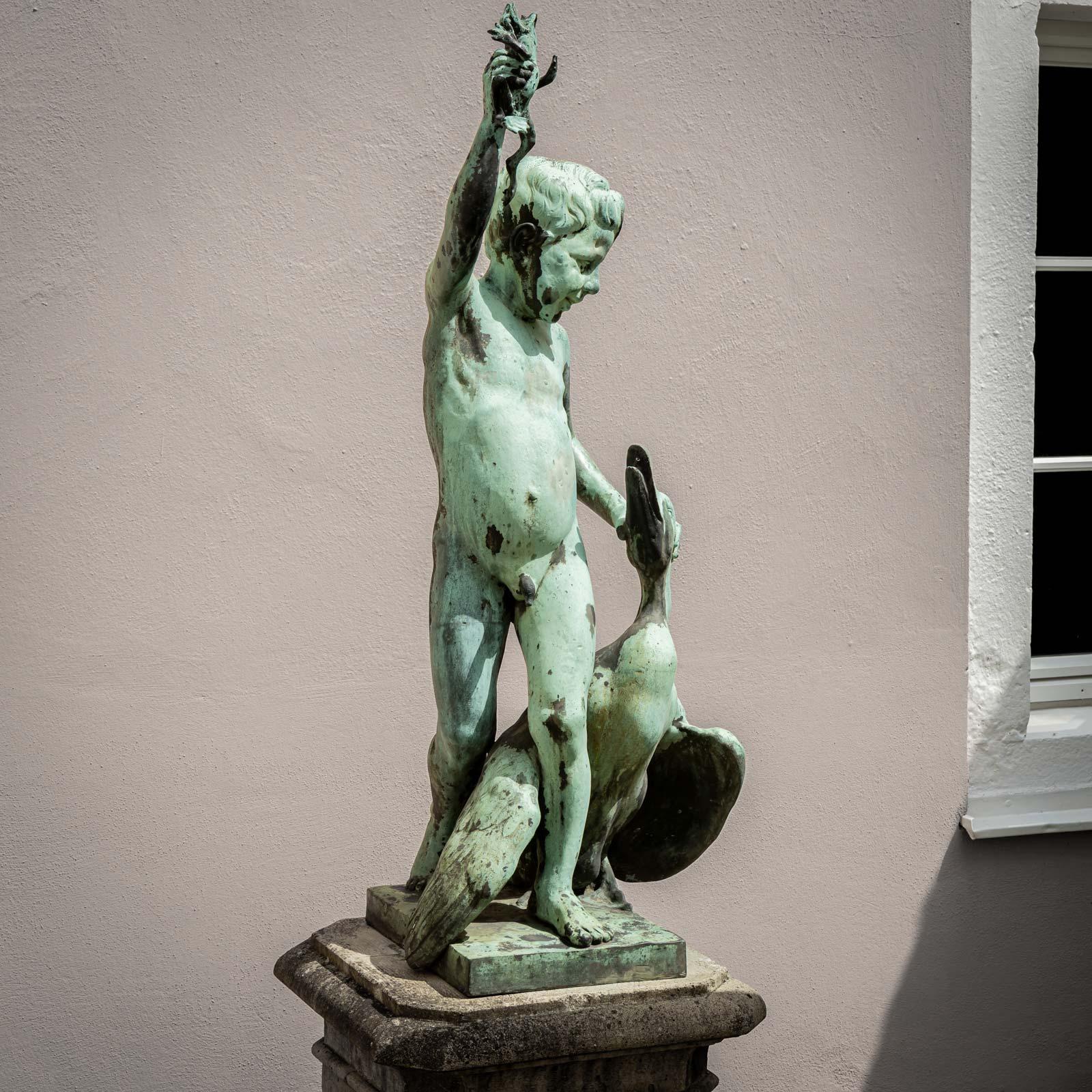 Figura in metallo fuso di un ragazzo con una rana in una mano e un'oca ai suoi piedi. La fontana esce dalla bocca della rana e dal becco dell'oca.