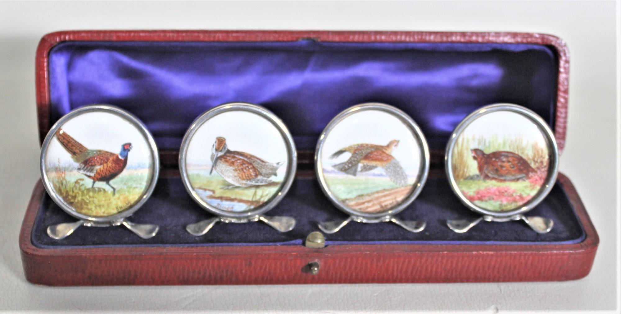 This set of four antique sterling silver place card or menu holders was made in Chester England in 1911 in an Edwardian style. Each of the card holders stand on a footed base with a large medallion with four different hand-painted game birds and