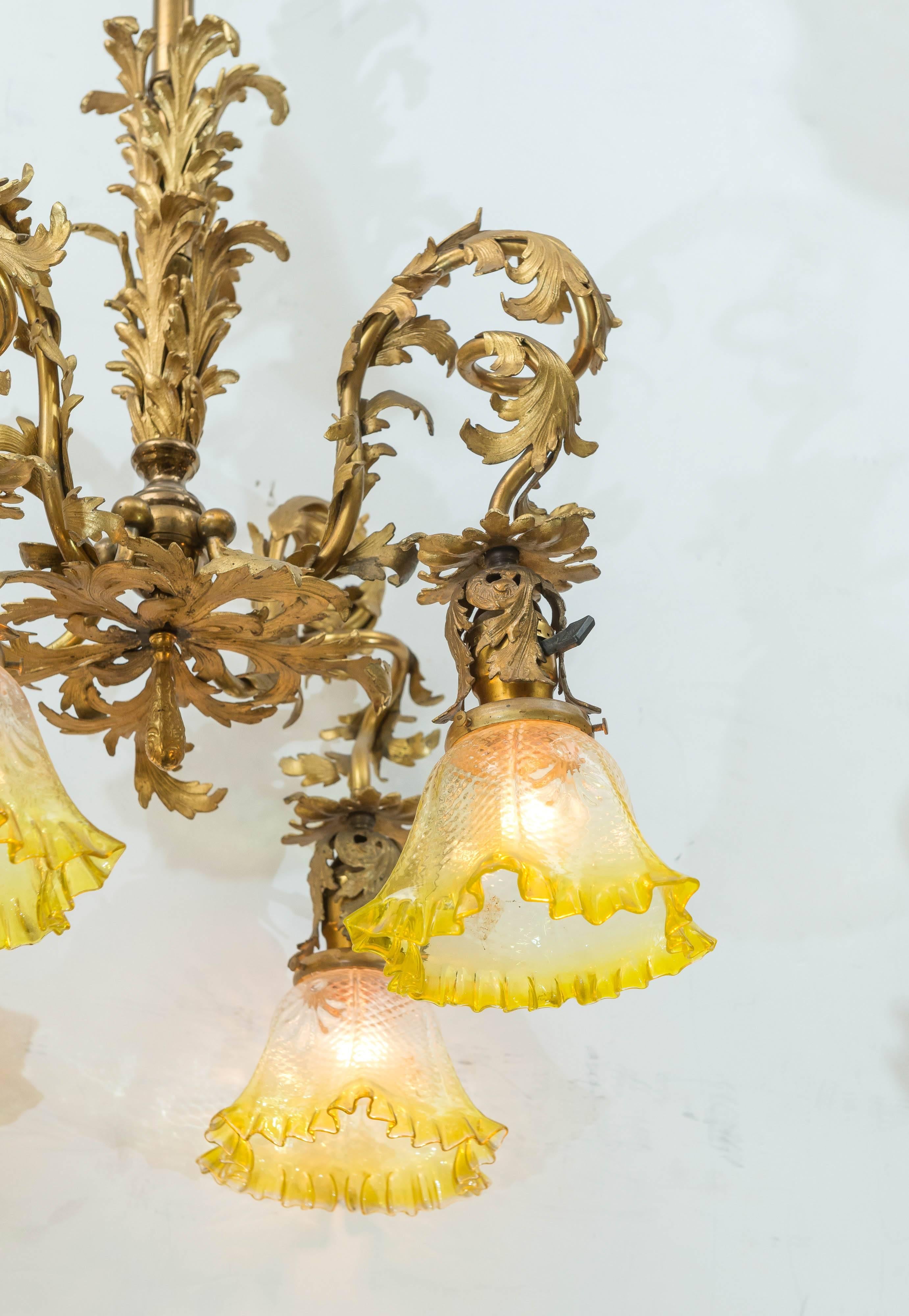 This American chandelier is done in the French taste and maintains it's original gilt finish. The glass shades are period and very unusual with highlighted frilly border. A perfect fit for this chandelier. Check out all the wonderful bronze work on