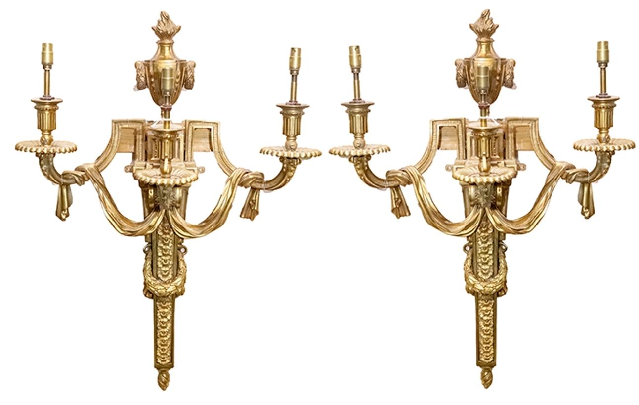 A good quality set of four classical Louis XVI style gilded ormolu three branch wall sconces, each having flaming urn finials, draping swags, graduating inset panels with flowers and a wreath around.